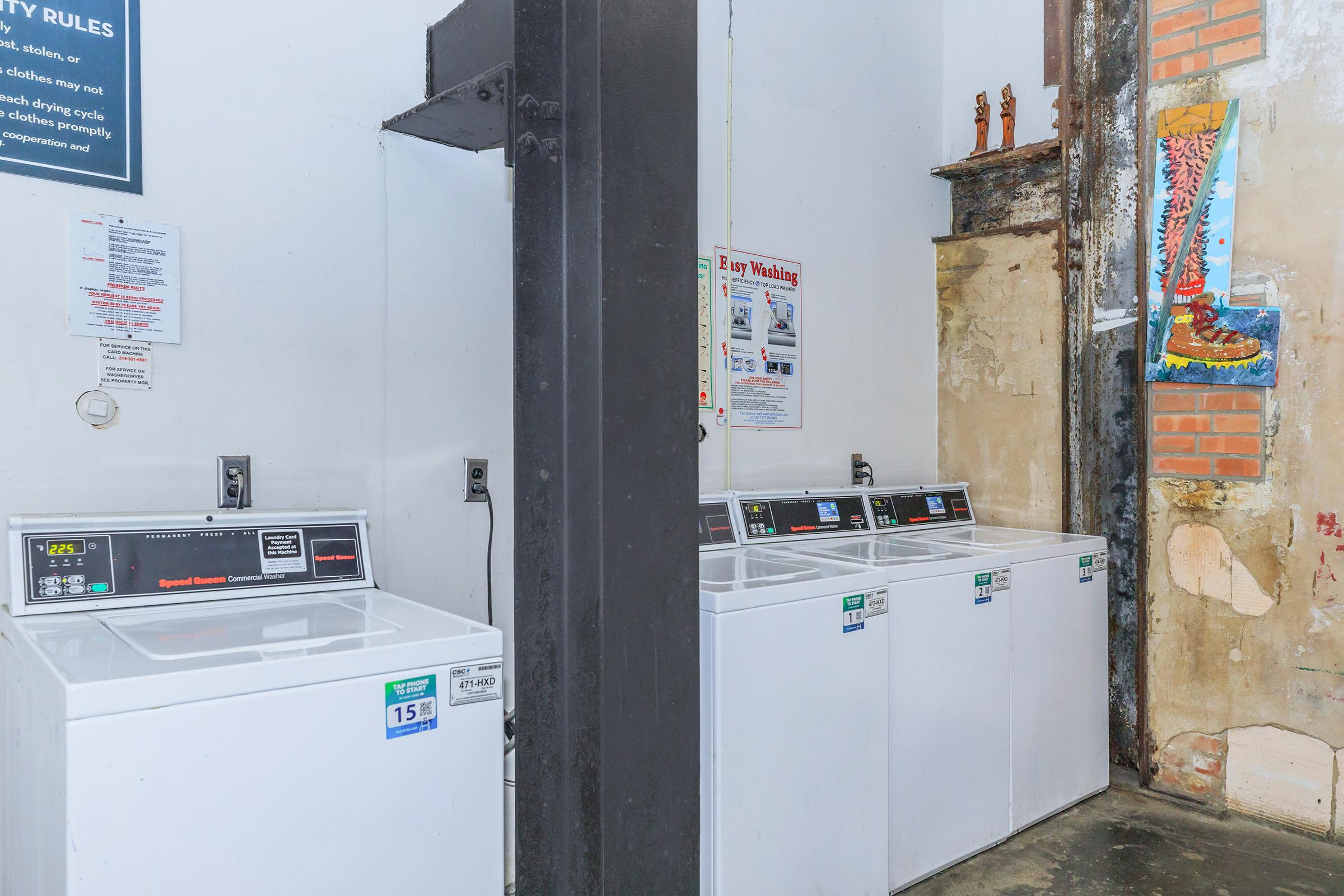 A laundry room featuring several white washing machines lined up against a wall, with informational posters about laundry rules and services displayed nearby. The walls show some wear, with exposed bricks and a colorful artwork of boots visible in the corner.
