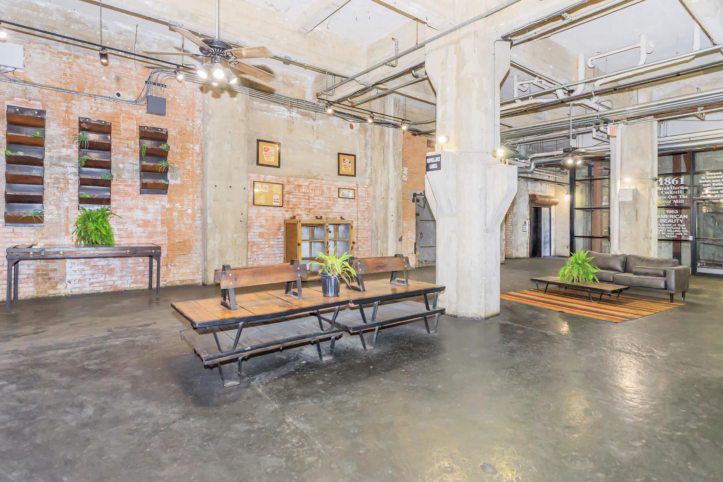 A spacious industrial-style lobby featuring exposed brick walls, concrete pillars, and modern furnishings. A long wooden table with chairs is paired with a cozy sofa. Decorative plants are present, and minimalist lighting adds ambiance. The area appears clean and well-maintained, suitable for gatherings or meetings.