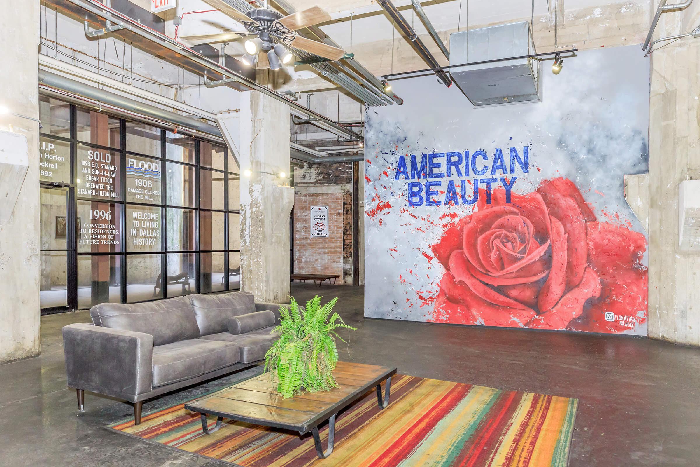 A modern interior space featuring a gray sofa and a wooden coffee table with a potted plant. The floor is covered with a colorful striped rug. A prominent mural on the wall displays the words "AMERICAN BEAUTY" alongside a large red rose, with exposed industrial elements and large windows in the background.