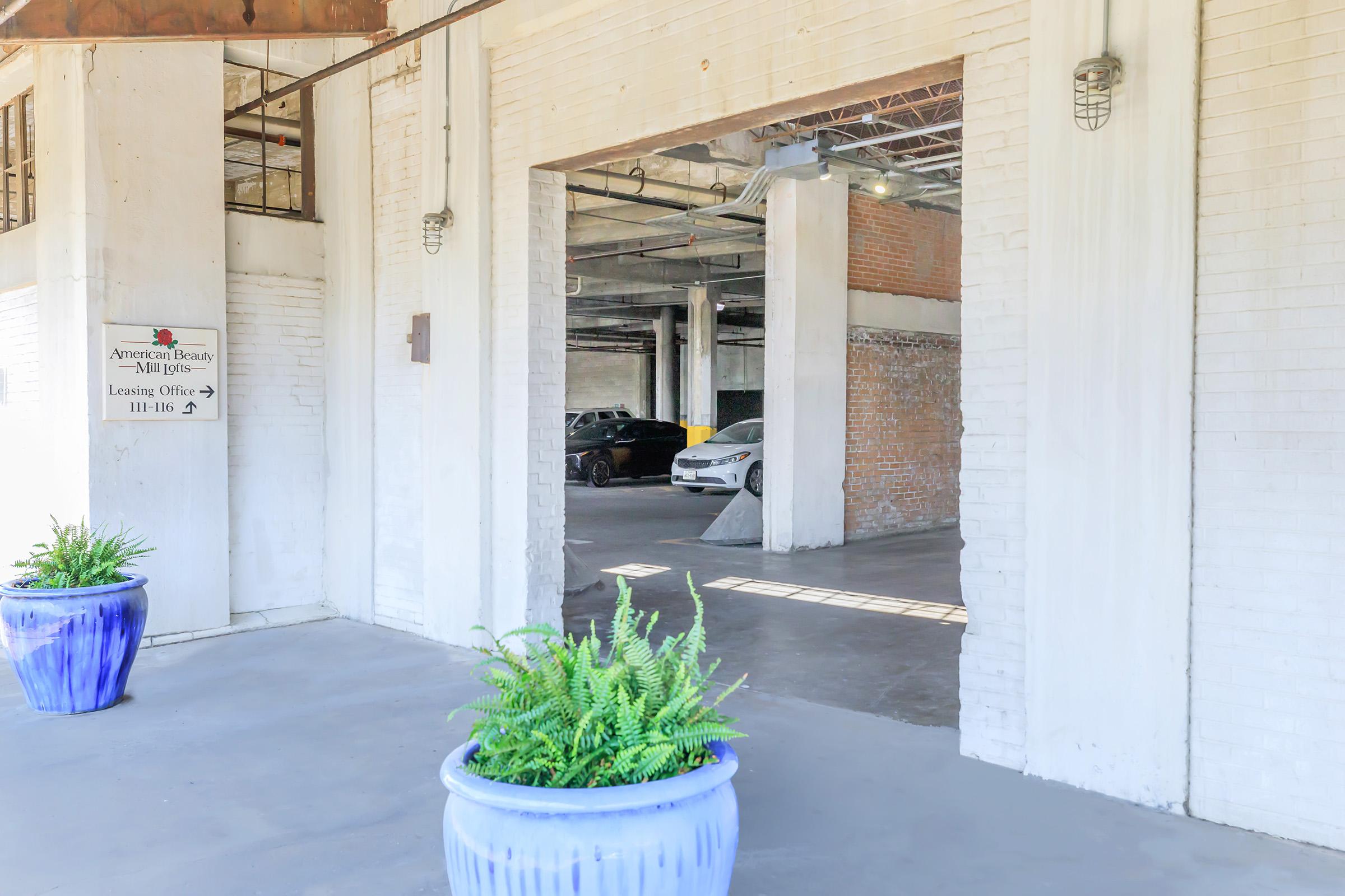 Entrance to a parking garage featuring a large open space with white walls and a brick accent wall. Two cars can be seen parked inside, and two blue planters with ferns add a touch of greenery at the entrance. A sign for "American Beauty Mill Office" is visible on the left side.