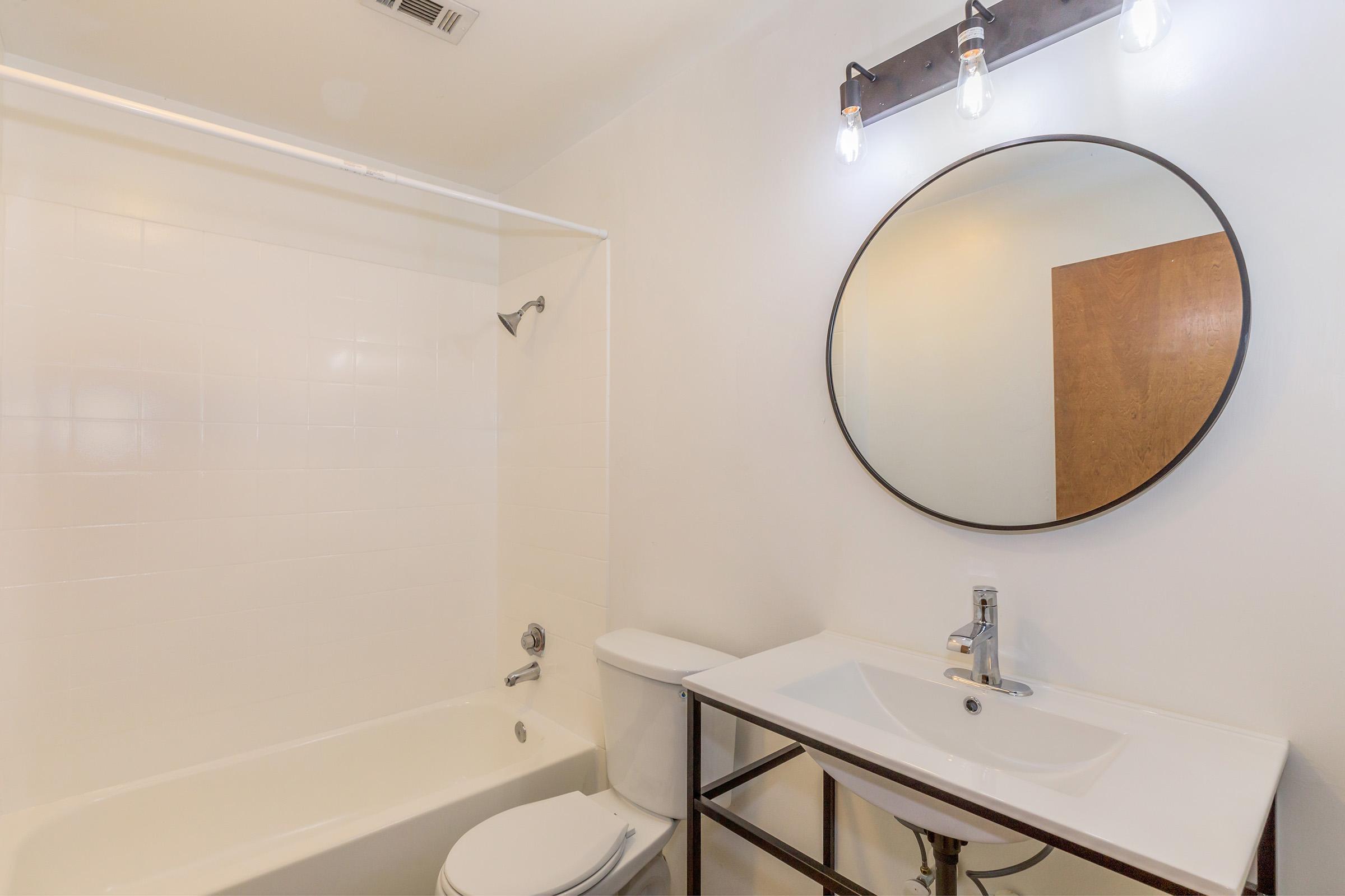 A modern bathroom featuring a white tub and shower combination, a circular mirror above a sleek sink with a contemporary faucet, and a dark metal shelf beneath the sink. The walls are painted white, and there is a wooden door visible in the background. Overhead lighting consists of exposed bulbs.