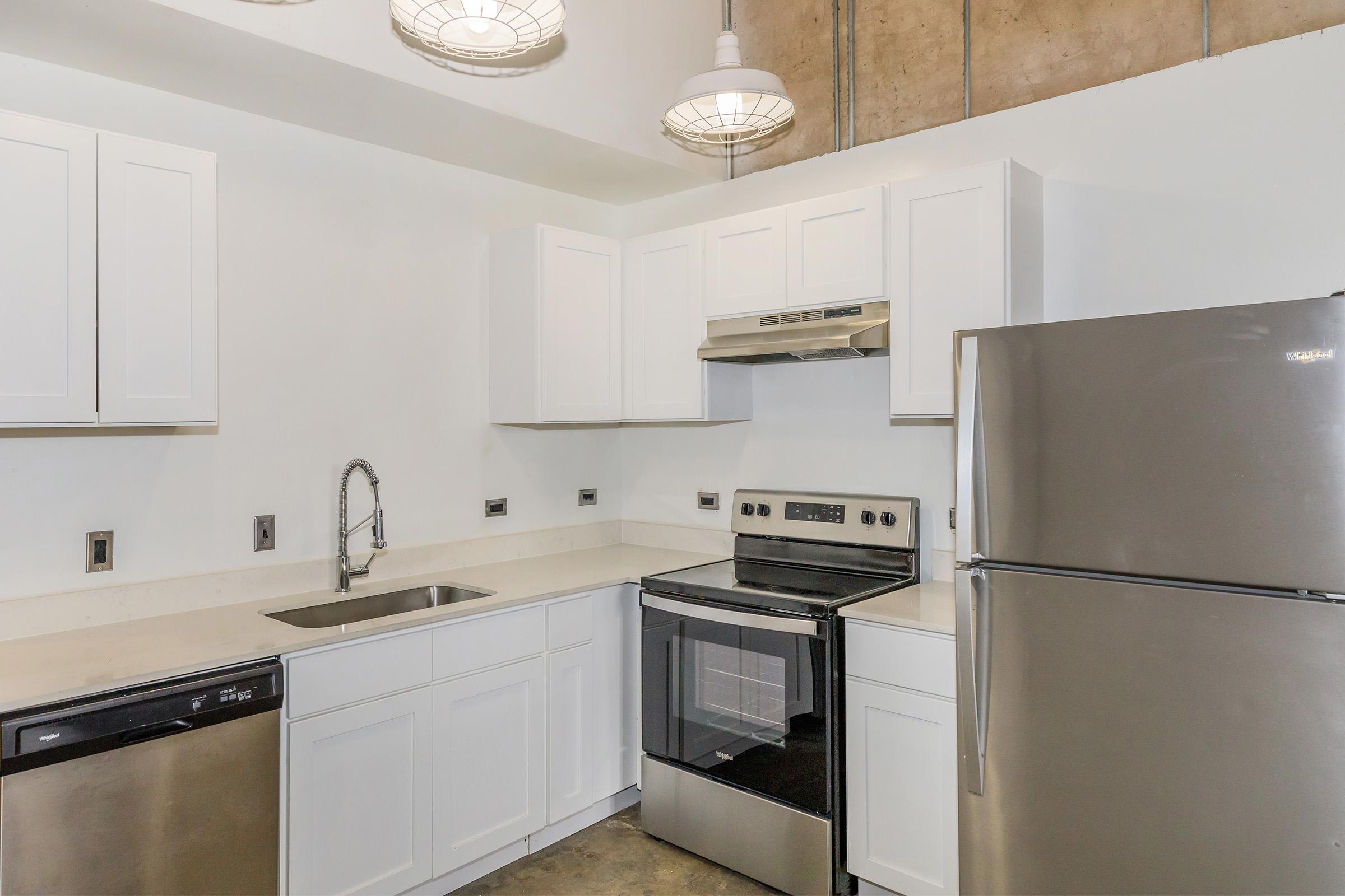 Modern kitchen featuring white cabinetry, stainless steel appliances including a refrigerator, oven, and dishwasher. The countertop is light-colored, and there are pendant lights hanging from the ceiling, with a concrete wall and flooring visible in the background.