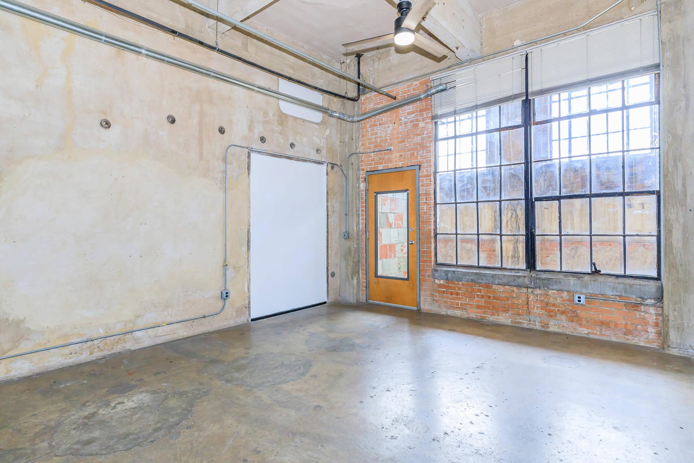 Interior of an industrial-style room featuring exposed brick walls, large windows with multiple panes, and a concrete floor. The space has a minimalist design with ceiling pipes and a ceiling fan, creating an open and airy atmosphere. A door is visible against the wall, leading to another area.
