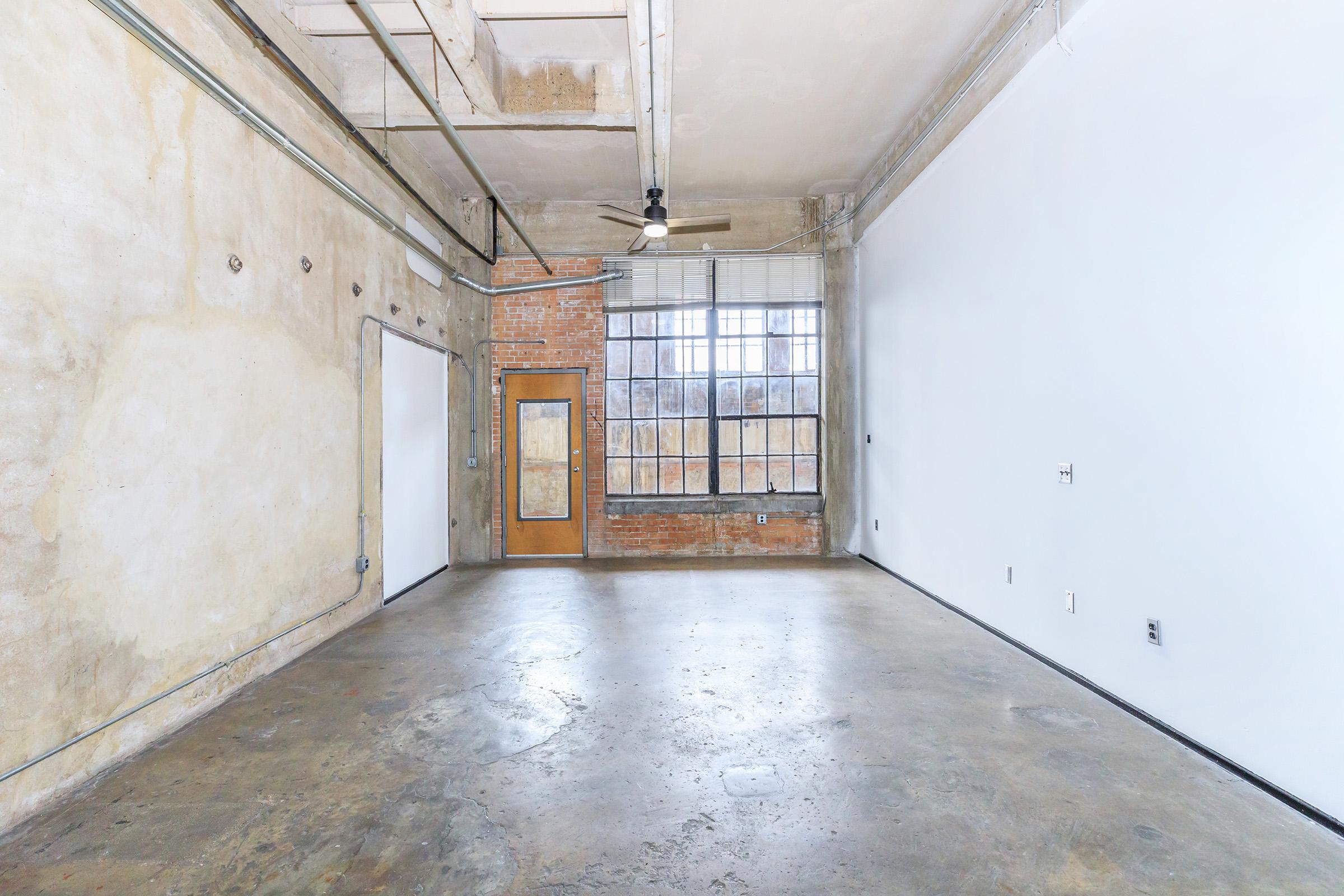 An empty industrial-style room with exposed brick walls and concrete flooring. Large windows let in natural light, and a ceiling fan is visible. One wall has a door, and the overall decor has a minimalist aesthetic. The space is open and versatile, suitable for various purposes.