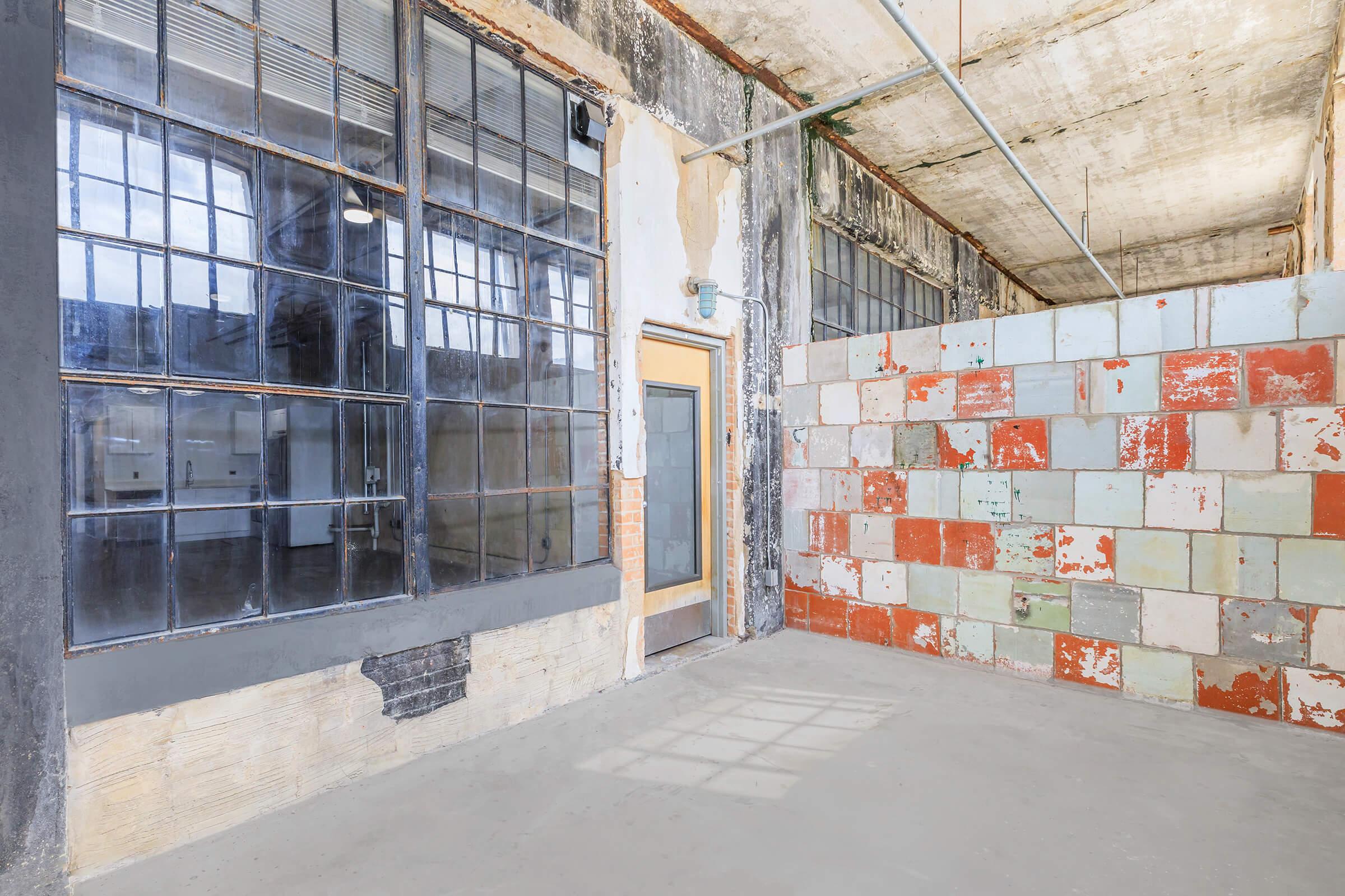 Spacious industrial interior featuring large windows and exposed concrete walls. A door on the left leads to another area, while a partition made of colorful tiles adds a unique design element. The floor is bare and the space is well-lit, highlighting the rustic atmosphere.