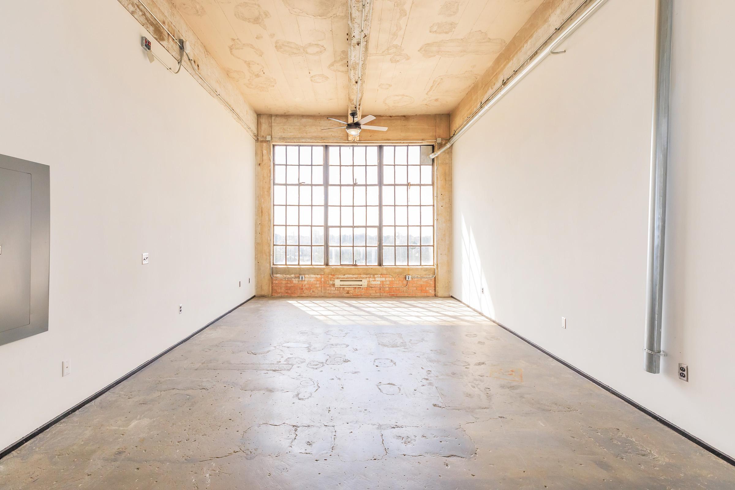 An empty room with a concrete floor and large industrial windows. Soft sunlight streams through the windows, casting shadows on the walls. The space features exposed brick, a ceiling fan, and minimal electrical fixtures, creating a bright and open atmosphere.