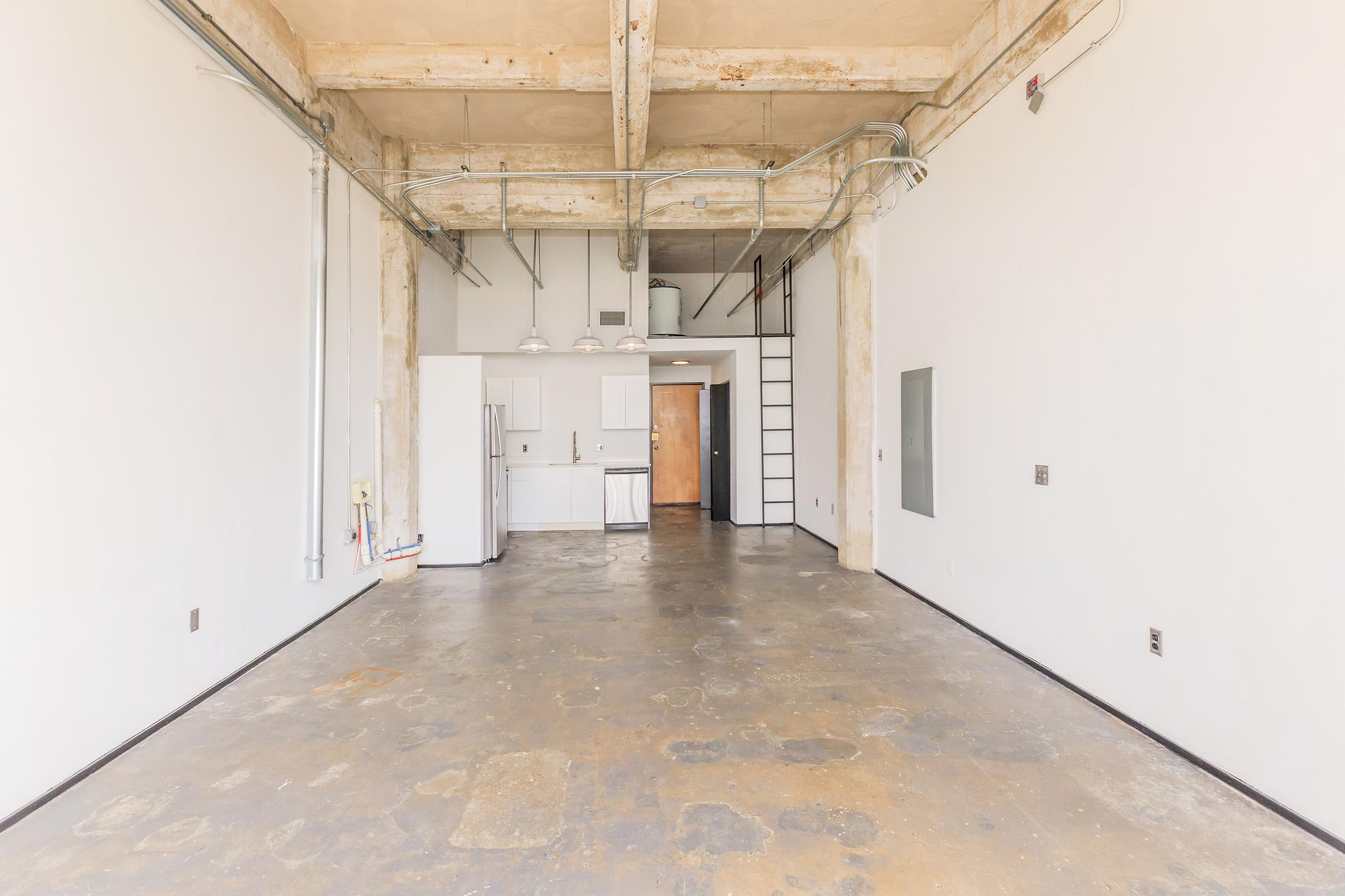 An empty, modern loft space featuring concrete floors and exposed beams. The room has large windows, minimal furniture, and an open layout with a kitchenette in the background. The walls are painted white, and industrial-style pipes are visible along the ceiling.