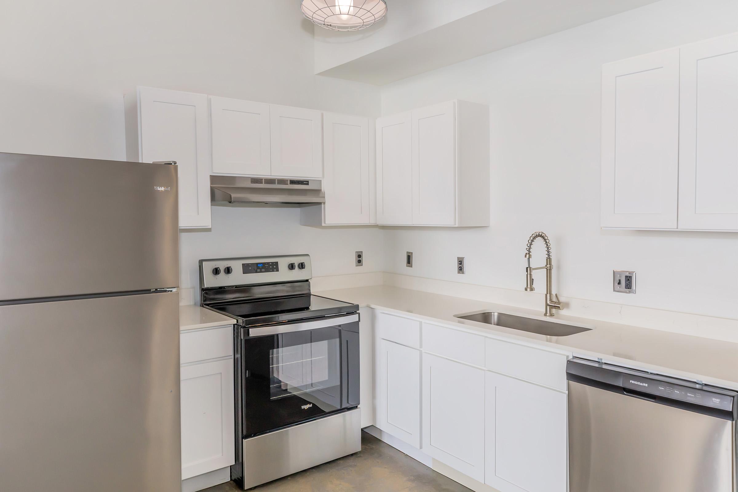 A modern kitchen featuring white cabinets, stainless steel appliances including a refrigerator, oven, and dishwasher, and a single-basin sink with a pull-down faucet. The space is well-lit with a pendant light and has a clean, contemporary design.