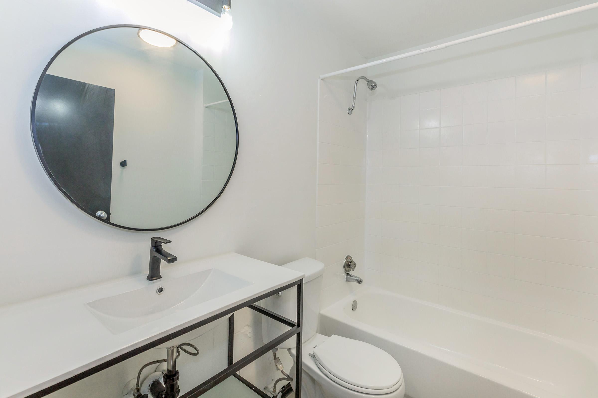 Modern bathroom featuring a round mirror above a sleek white sink with a black faucet. A glass shelving unit complements the minimalist design. On the right, a white bathtub with a showerhead is visible against white tiled walls. The overall aesthetic is clean and contemporary, with bright lighting.