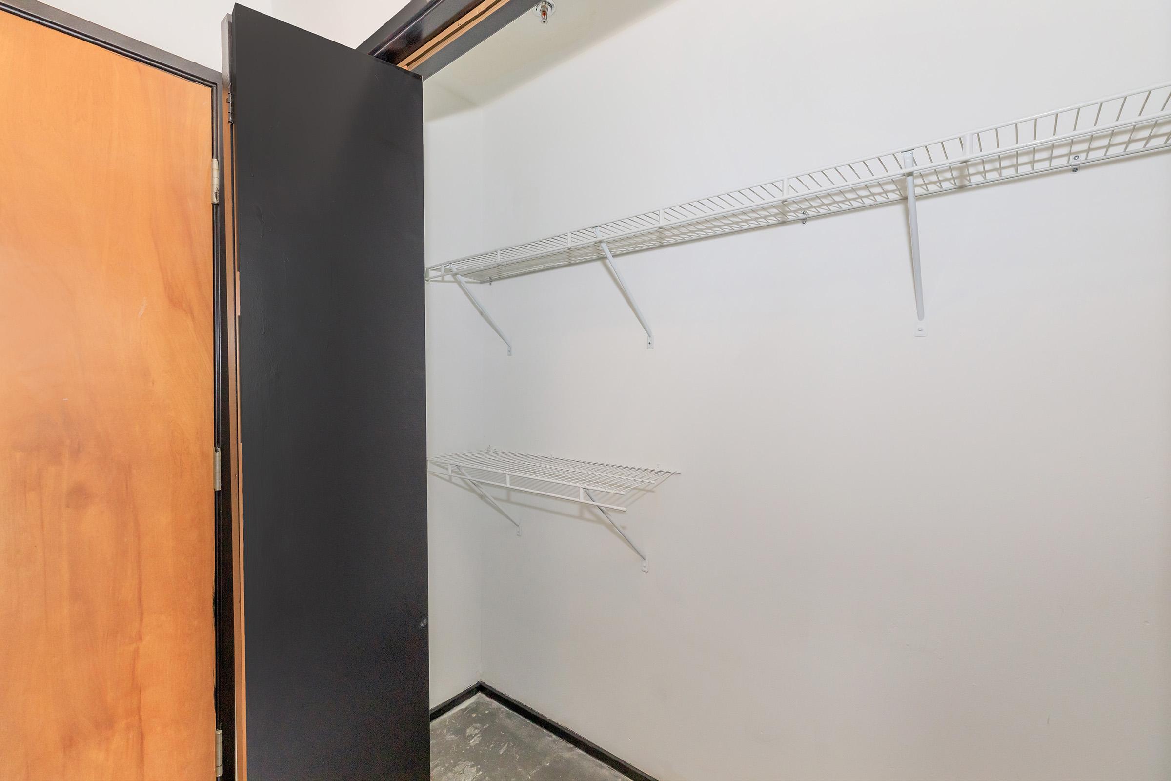 Interior of a closet featuring bare walls and two metal wire shelves. The closet has a black door partially open, revealing a minimalist and empty space without any additional storage items or decorations. The floor is concrete, and the overall appearance is clean and simple.