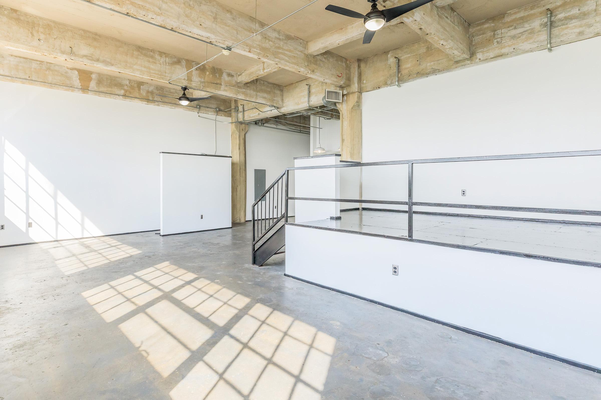 A spacious, industrial-style interior featuring exposed concrete beams and large windows casting shadows on the floor. The room includes a small staircase leading to an elevated platform with a railing, and white walls that enhance the bright, open atmosphere. Minimalist design with plenty of natural light.