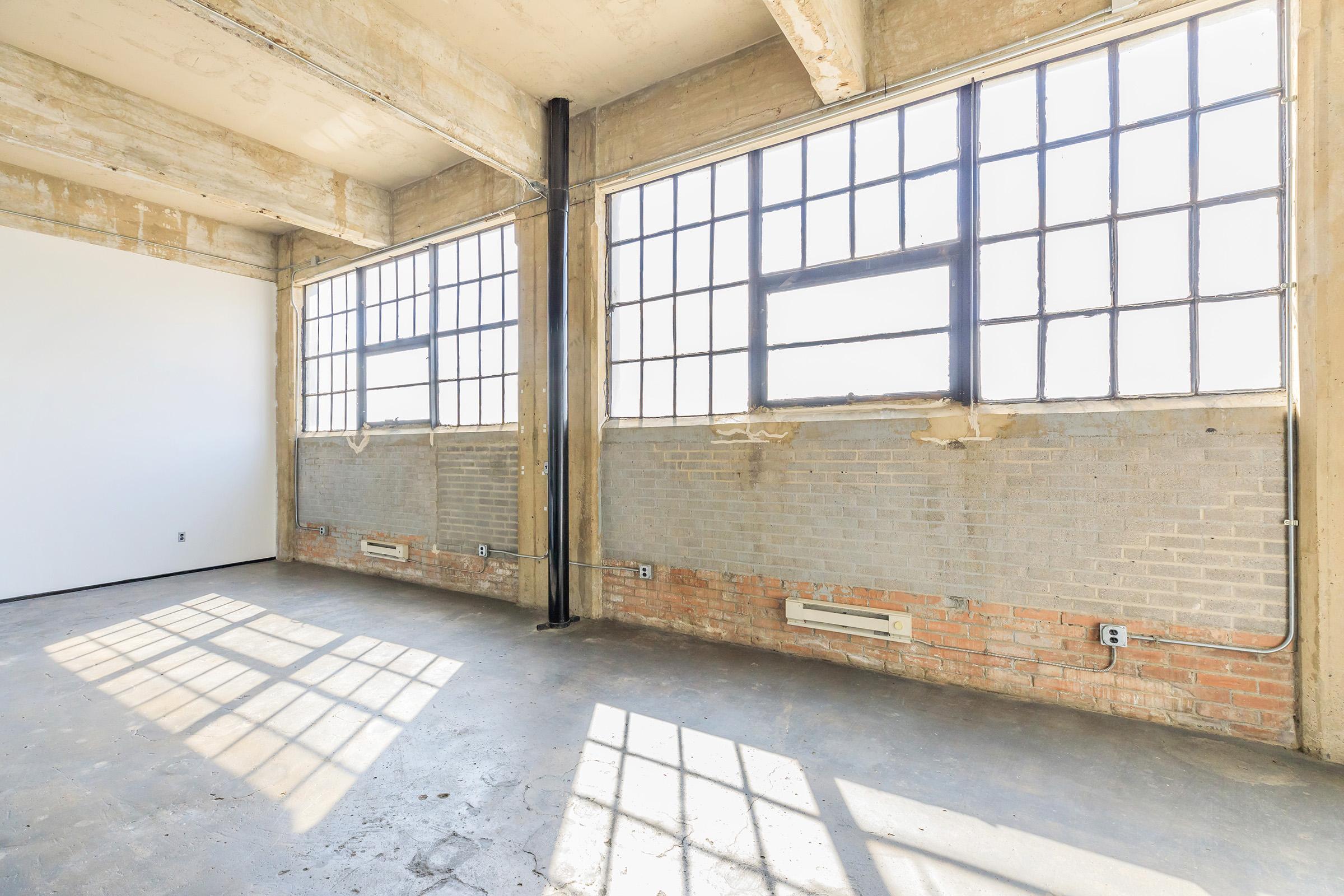 A spacious, empty room featuring large industrial-style windows with metal frames. The walls are a mix of exposed brick and smooth concrete, with visible electrical outlets and pipes. Sunlight casts shadows on the concrete floor, highlighting the room's open and airy atmosphere.