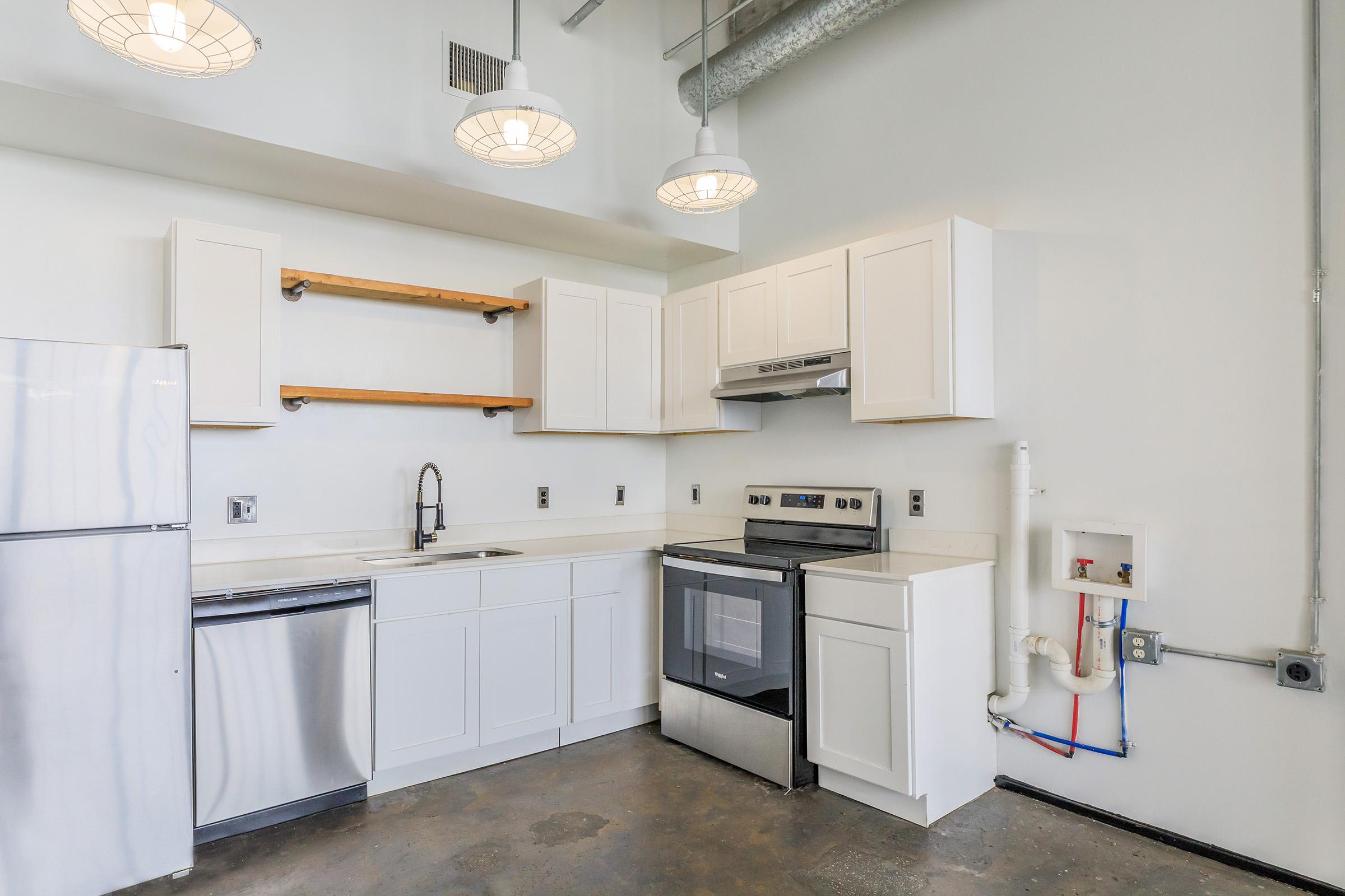 Modern kitchen with white cabinetry, stainless steel appliances including a refrigerator, dishwasher, and oven. Features a simple countertop and minimalist design with open shelving. The walls are light-colored and the floor is concrete. Three pendant lights hang above the kitchen area.
