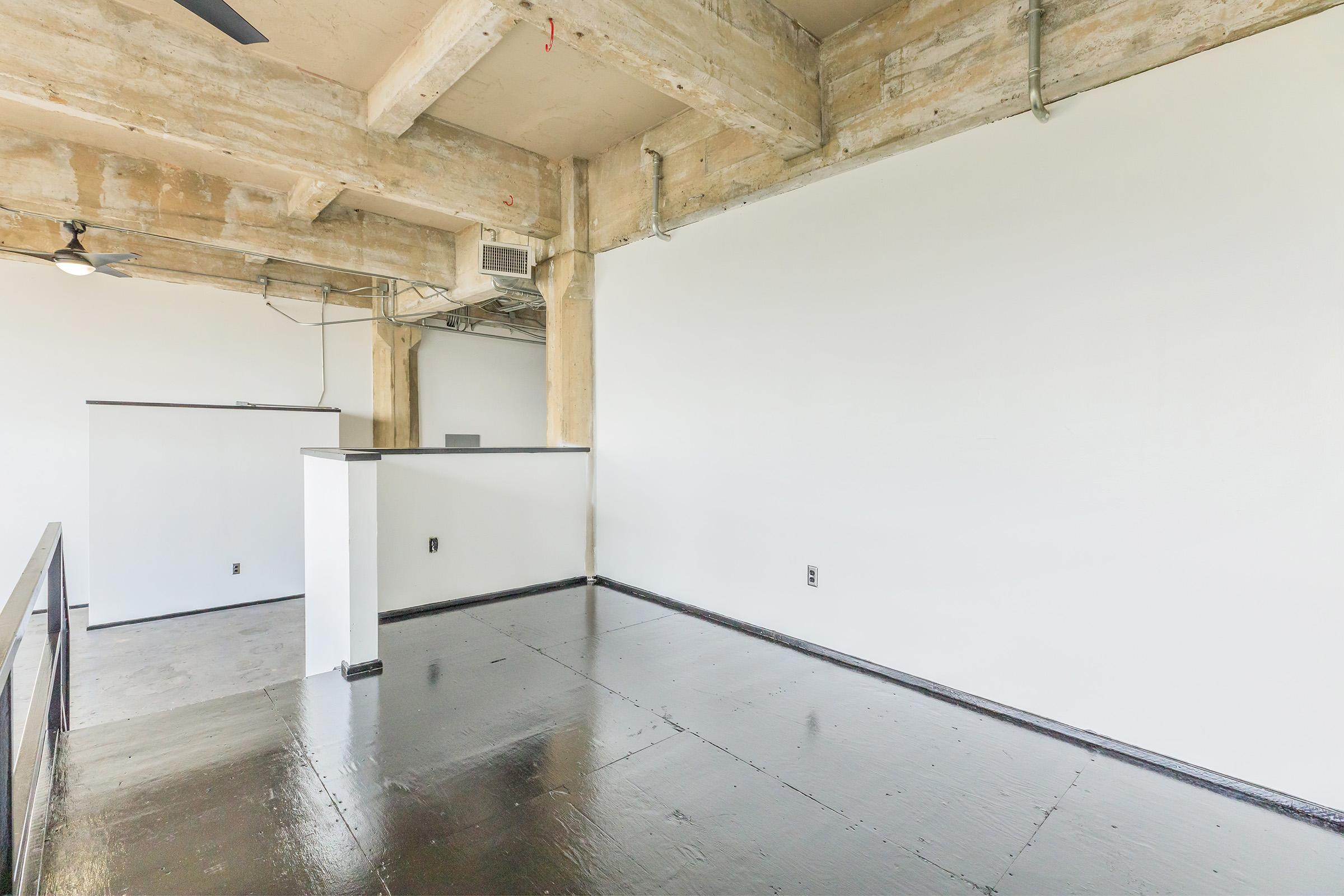 A spacious, minimalist interior of a room featuring concrete walls and exposed wooden beams. The floor is glossy and black, while the walls are painted white. There’s a small partition and an air conditioning unit visible, contributing to the open and modern aesthetic. The overall look is industrial yet clean.