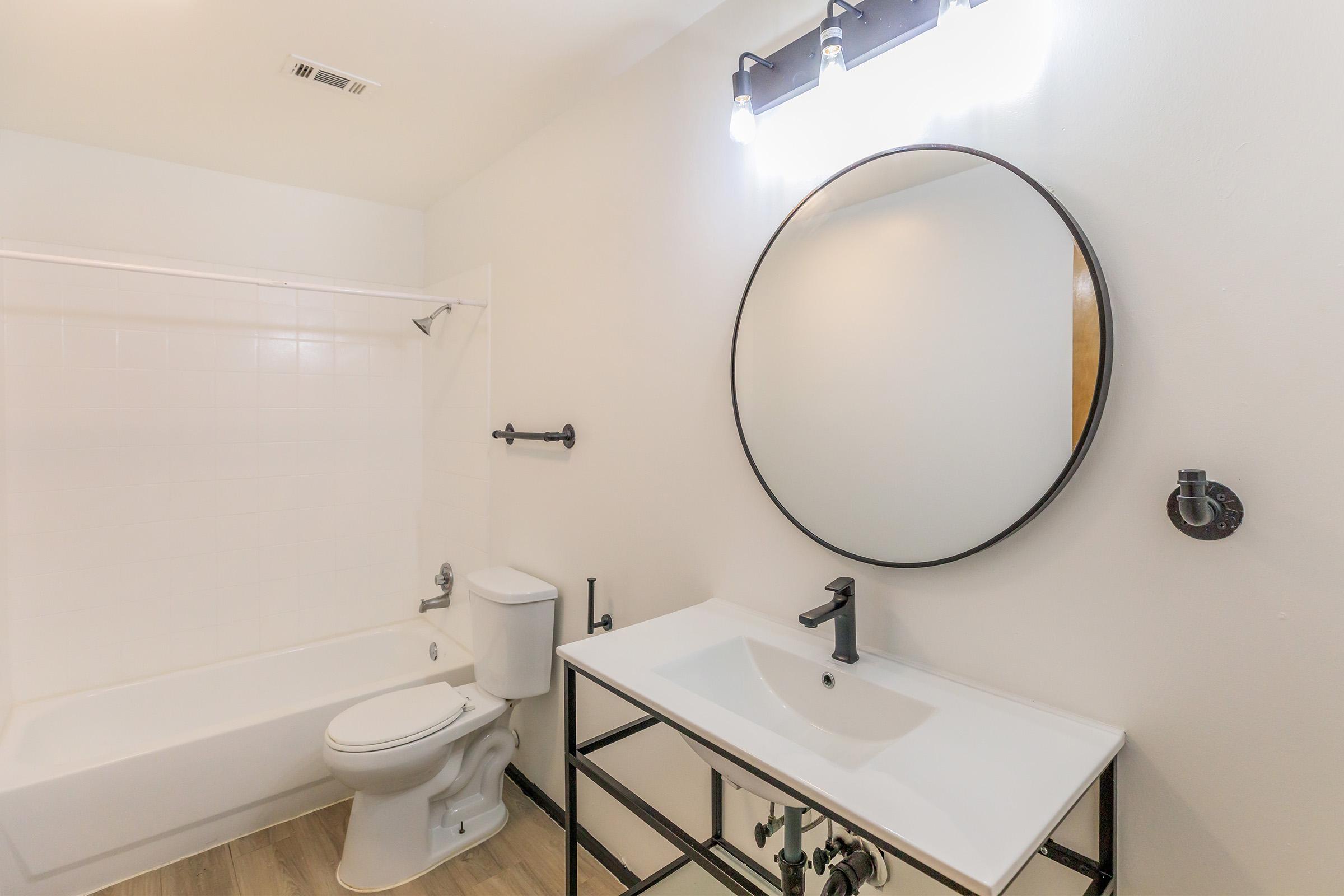 A modern bathroom featuring a circular mirror above a sleek white sink, a black faucet, and a black metal base. The space includes a toilet and a bathtub with a shower. The walls are painted white, and there are bright overhead lights illuminating the area. Floor tiles are light-colored wood.