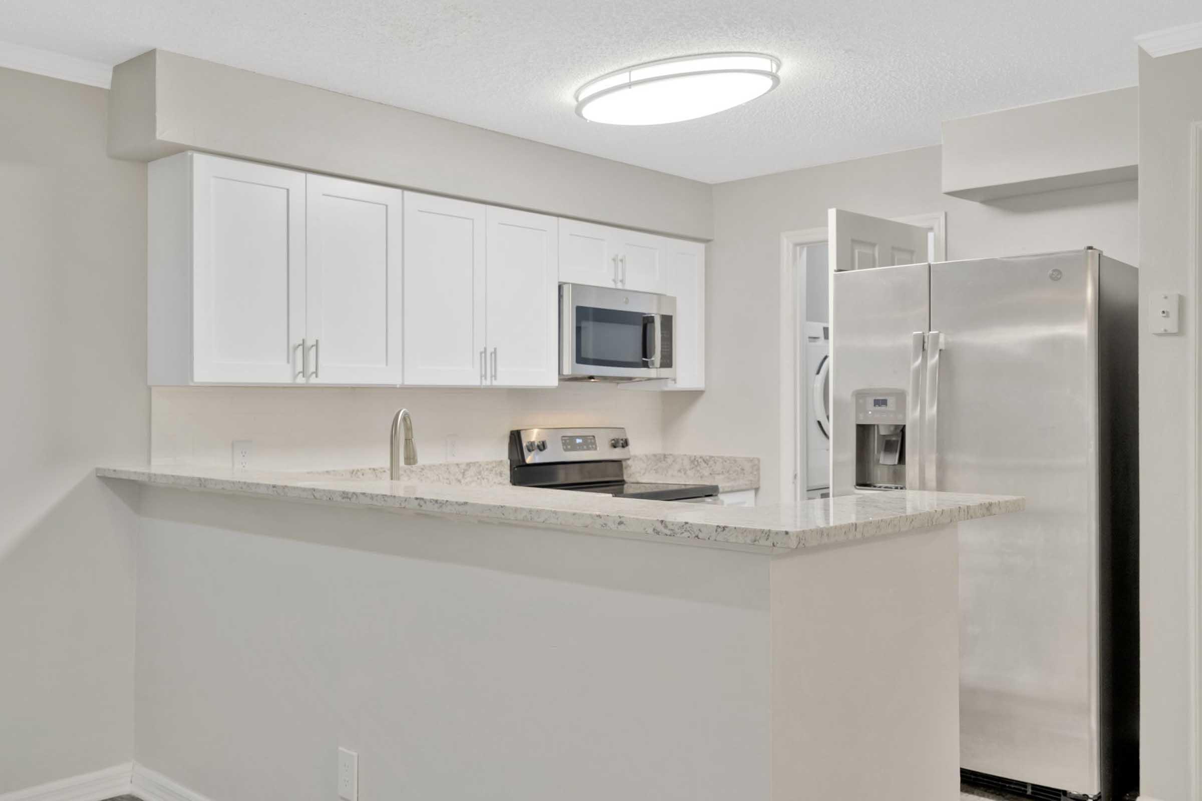 Contemporary kitchen featuring a light-colored granite countertop, white cabinetry, a stainless steel refrigerator, a built-in microwave, and a modern stove. A circular ceiling light illuminates the space, creating a bright and inviting atmosphere.