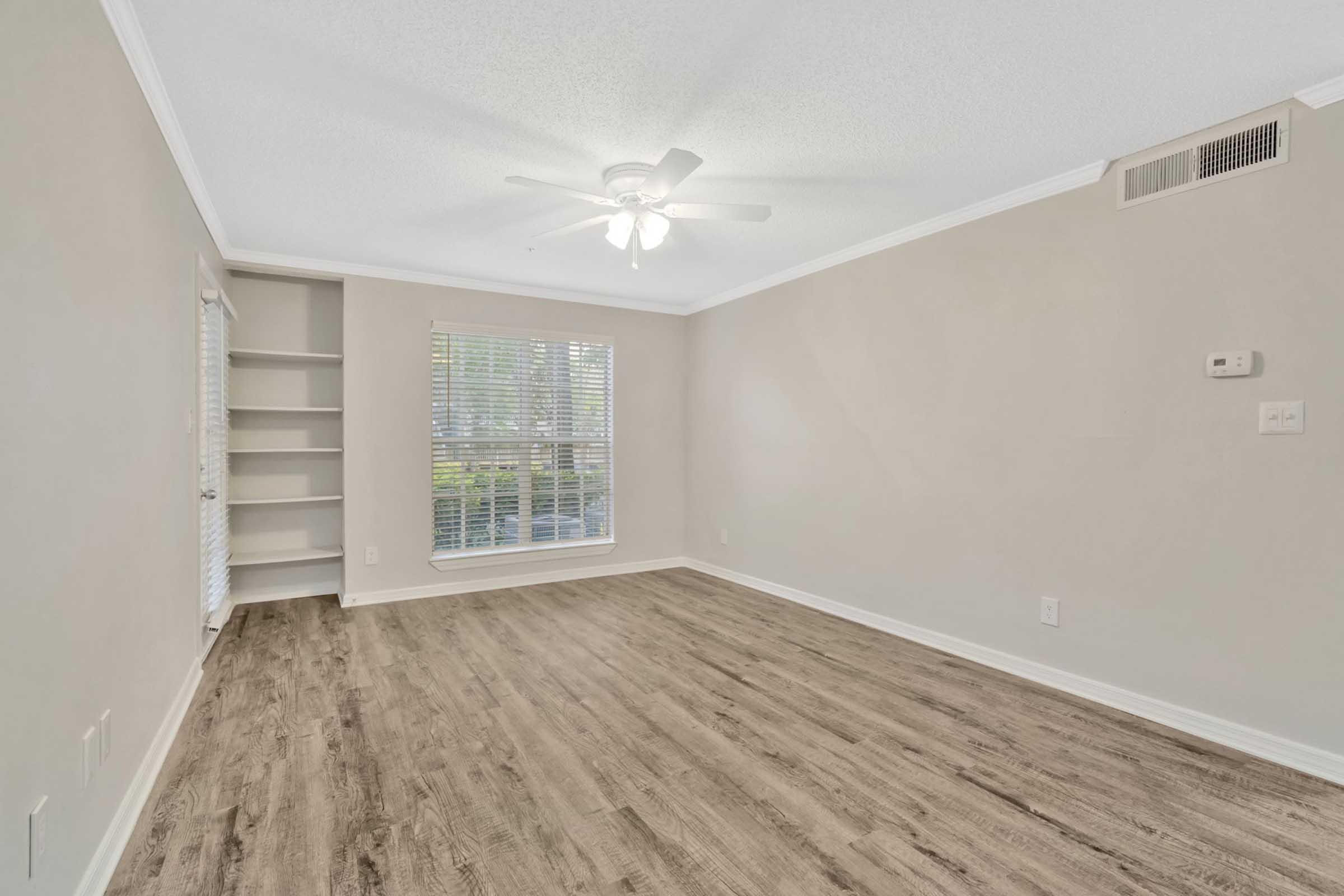A spacious, empty living room with light-colored walls, a ceiling fan, and laminate flooring. A large window lets in natural light, and there are built-in shelves on one side. The room is well-lit and invites a welcoming atmosphere.