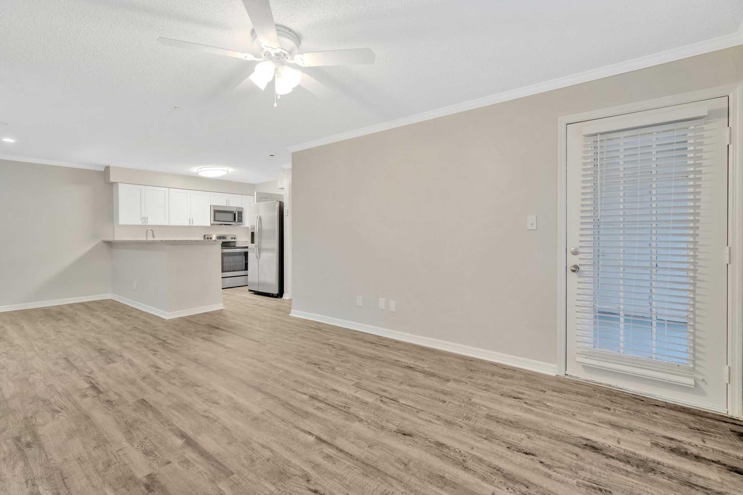 Interior of a modern apartment featuring an open floor plan. The living area has light wood-like flooring, a ceiling fan, and a doorway with blinds leading outside. The kitchen, visible in the background, has white cabinets and stainless steel appliances.