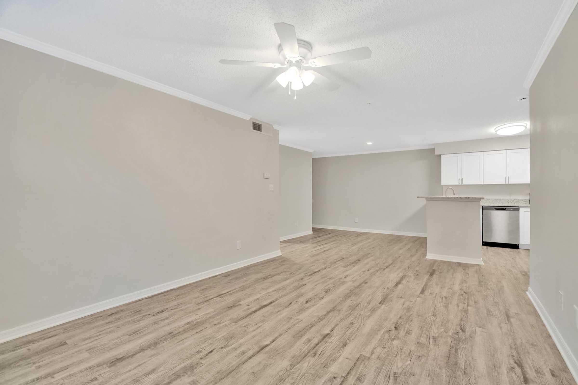 A spacious, empty room featuring light-colored walls, a ceiling fan, and hardwood-style flooring. In the background, there is a kitchen area with white cabinets and stainless steel appliances. The open layout gives a modern feel with ample natural light.
