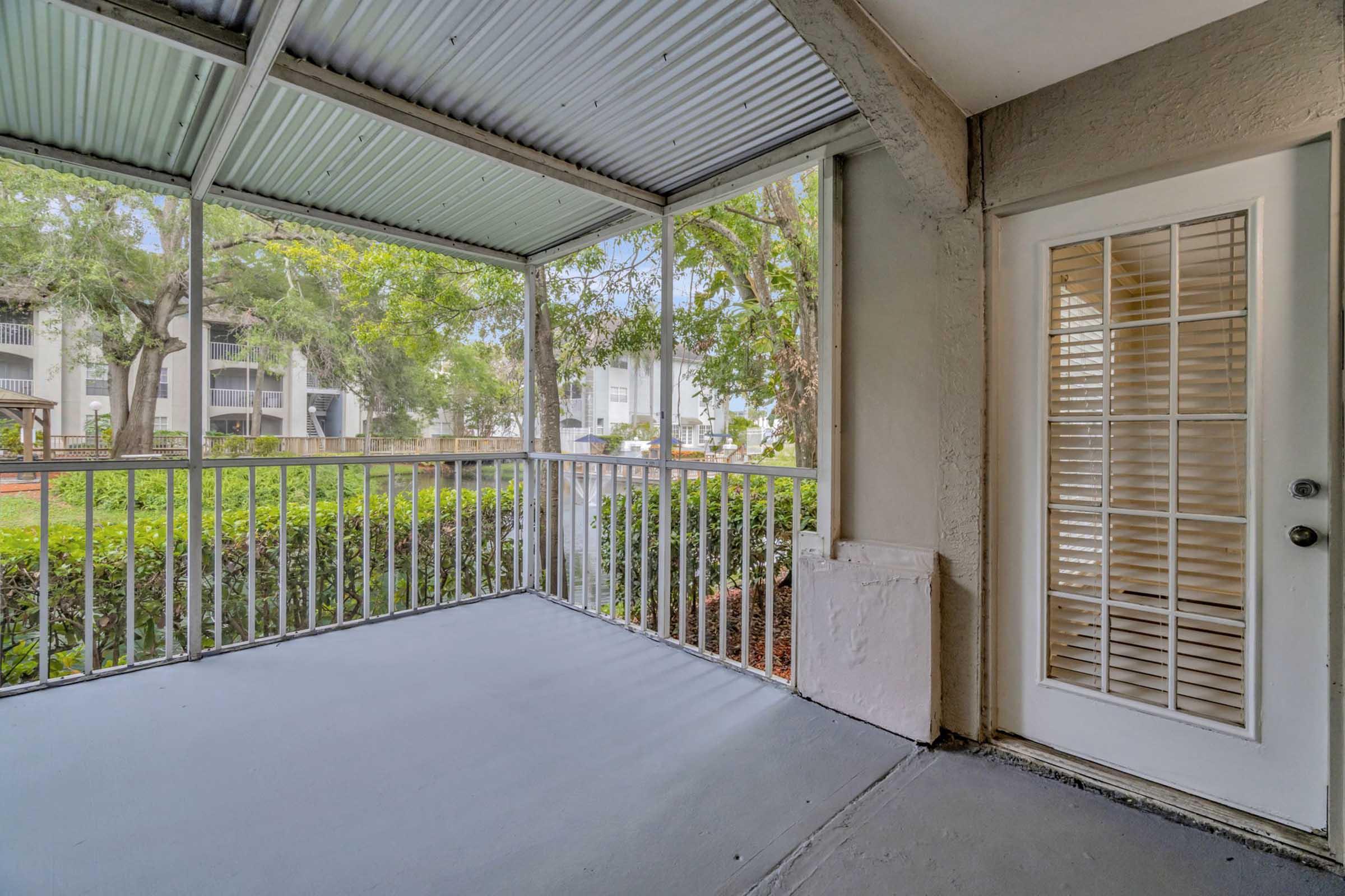 A screened-in porch with a gray floor, featuring a white door with shutters, surrounded by trees and greenery. The space is well-lit with natural light coming through the screens, providing a peaceful outdoor view.