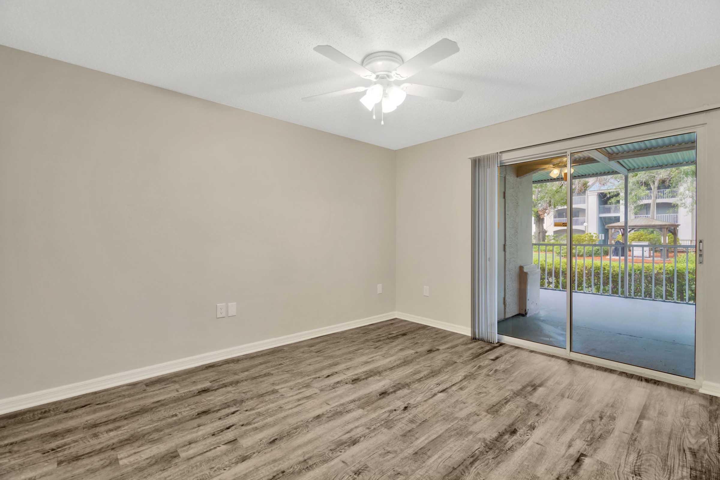 A spacious, well-lit room featuring a ceiling fan, with light-colored walls and hardwood-style flooring. A sliding glass door leads to a porch area with outdoor views, creating a welcoming and airy atmosphere.