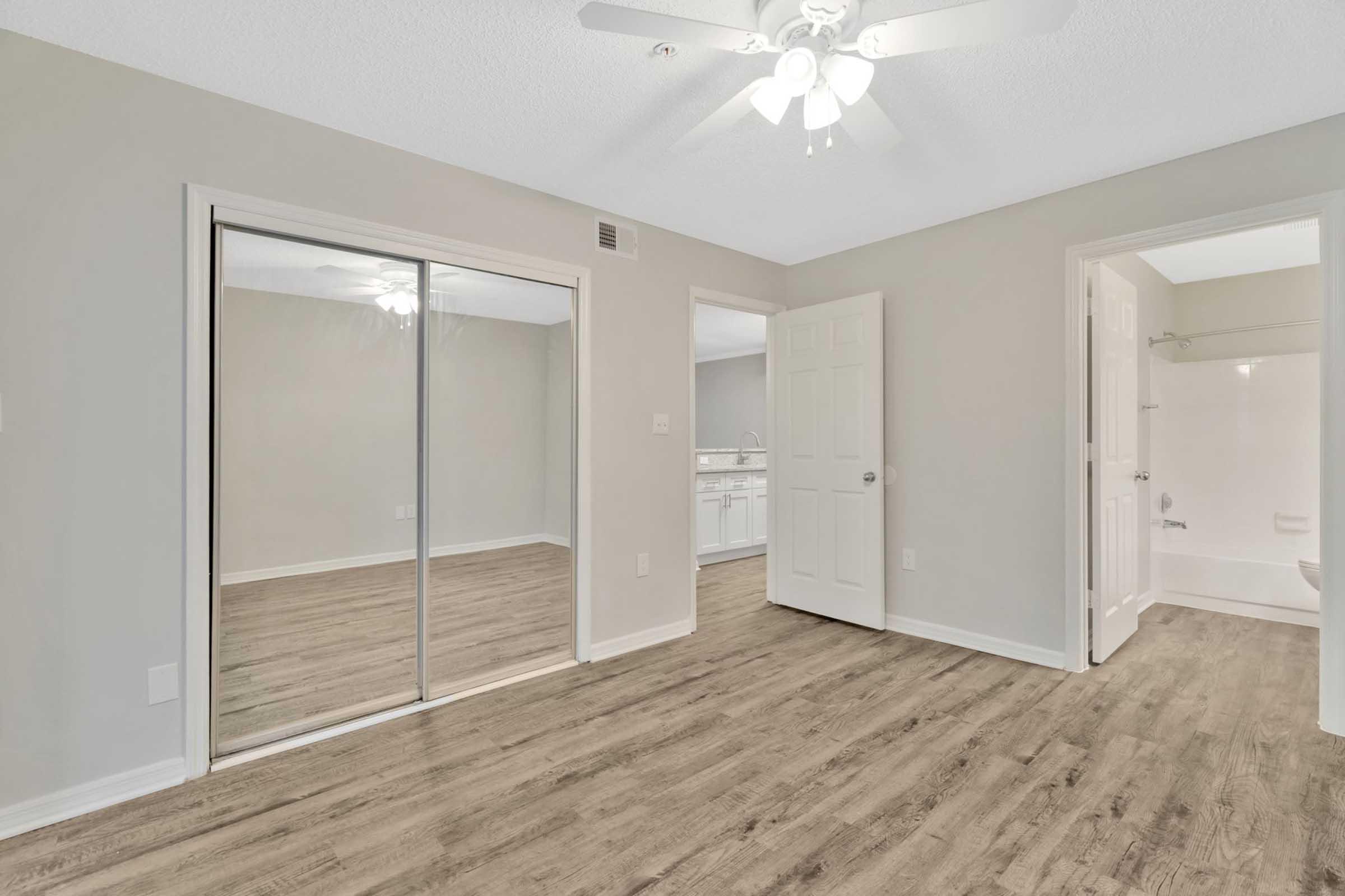 A spacious room with light gray walls, ample natural light, and a ceiling fan. There is a mirrored closet on one wall, and an open door leading to a bathroom area visible in the background. The flooring is a warm wood laminate, creating a cozy atmosphere.