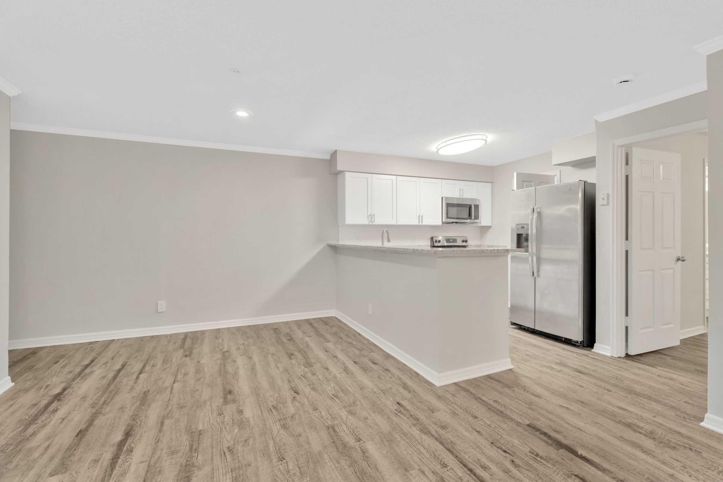 A spacious, modern kitchen and living area featuring light gray walls, wood-like flooring, and a kitchenette with white cabinets, a microwave, and stainless steel appliances. The area is well-lit with a circular ceiling light.