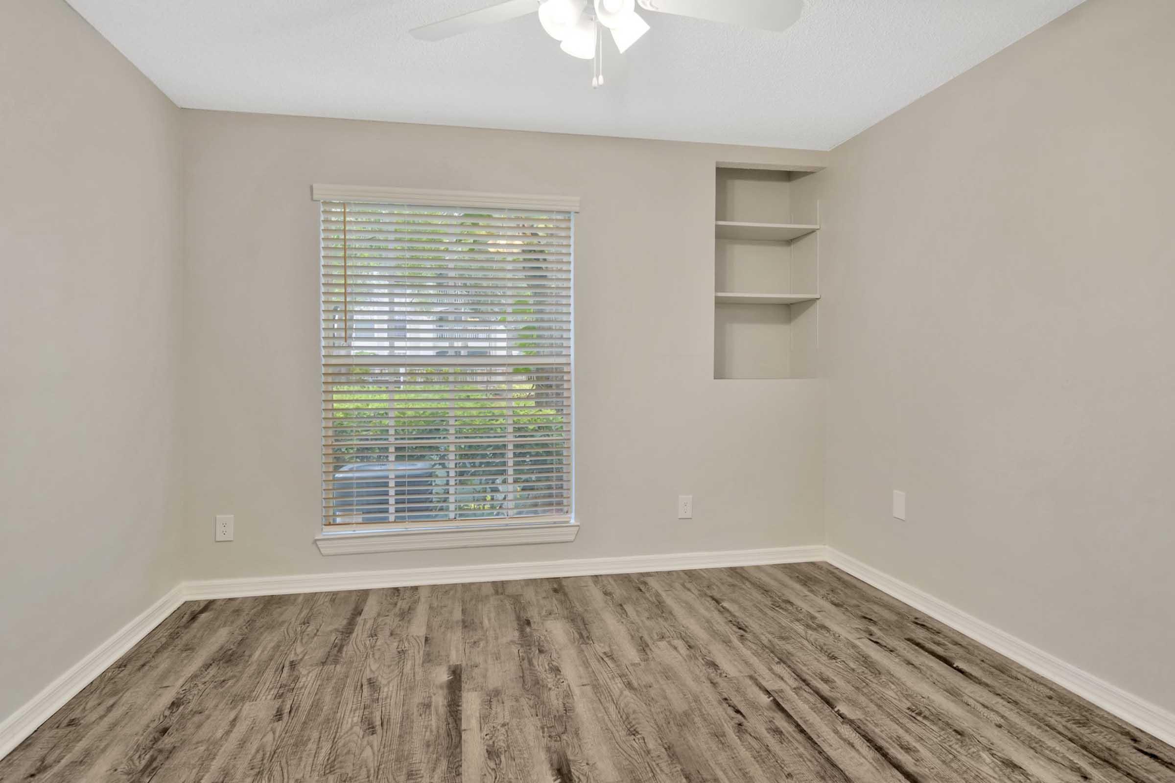 A well-lit empty room with light gray walls, featuring a window with blinds, a ceiling fan, and a wooden floor. There are built-in shelves on one wall, and the space is free of furniture, creating a clean and open atmosphere.