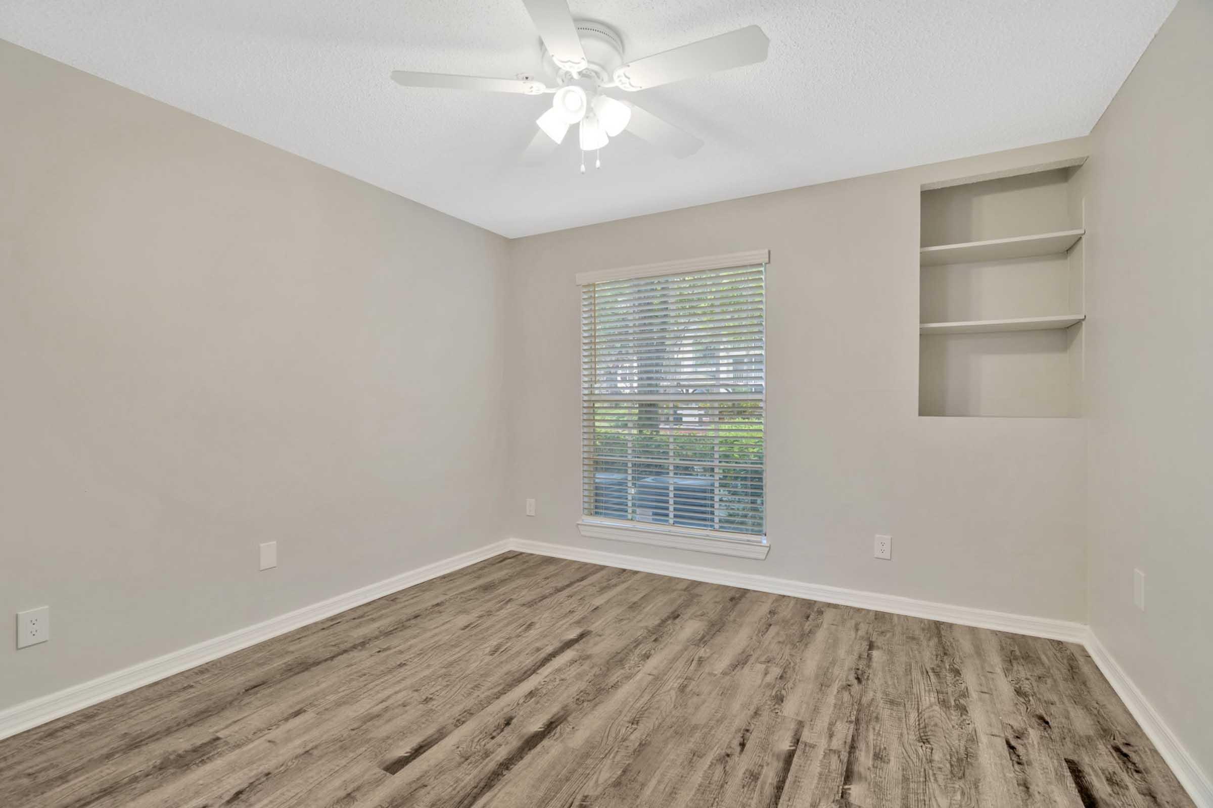 Empty room with light-colored walls, a ceiling fan, and a window with closed blinds. There is a built-in shelf on the right wall. The floor is composed of light wood laminate, creating a bright and spacious atmosphere.