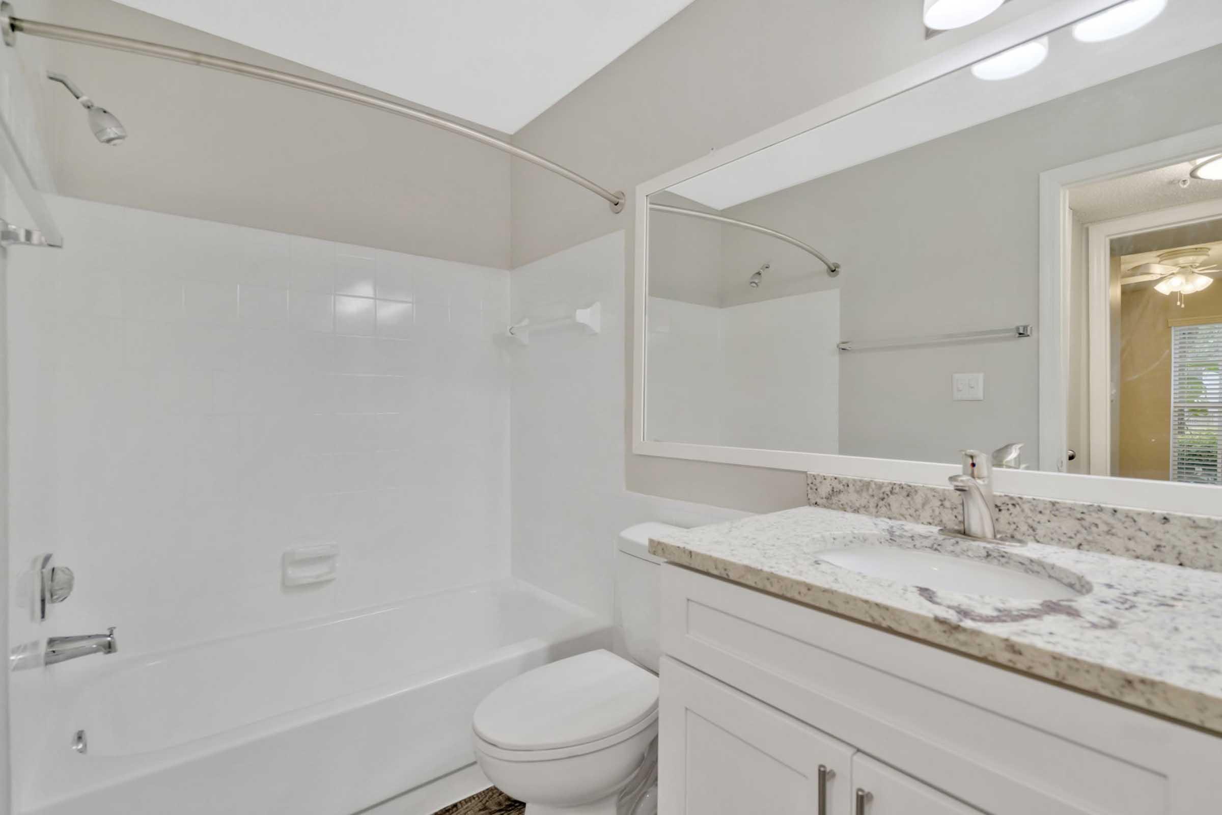 A clean and modern bathroom featuring a white bathtub with a shower, a large mirror above a granite countertop sink, and a toilet. The walls are painted in a light color, creating a spacious feel, and there is a glimpse of a light fixture and window in the background.
