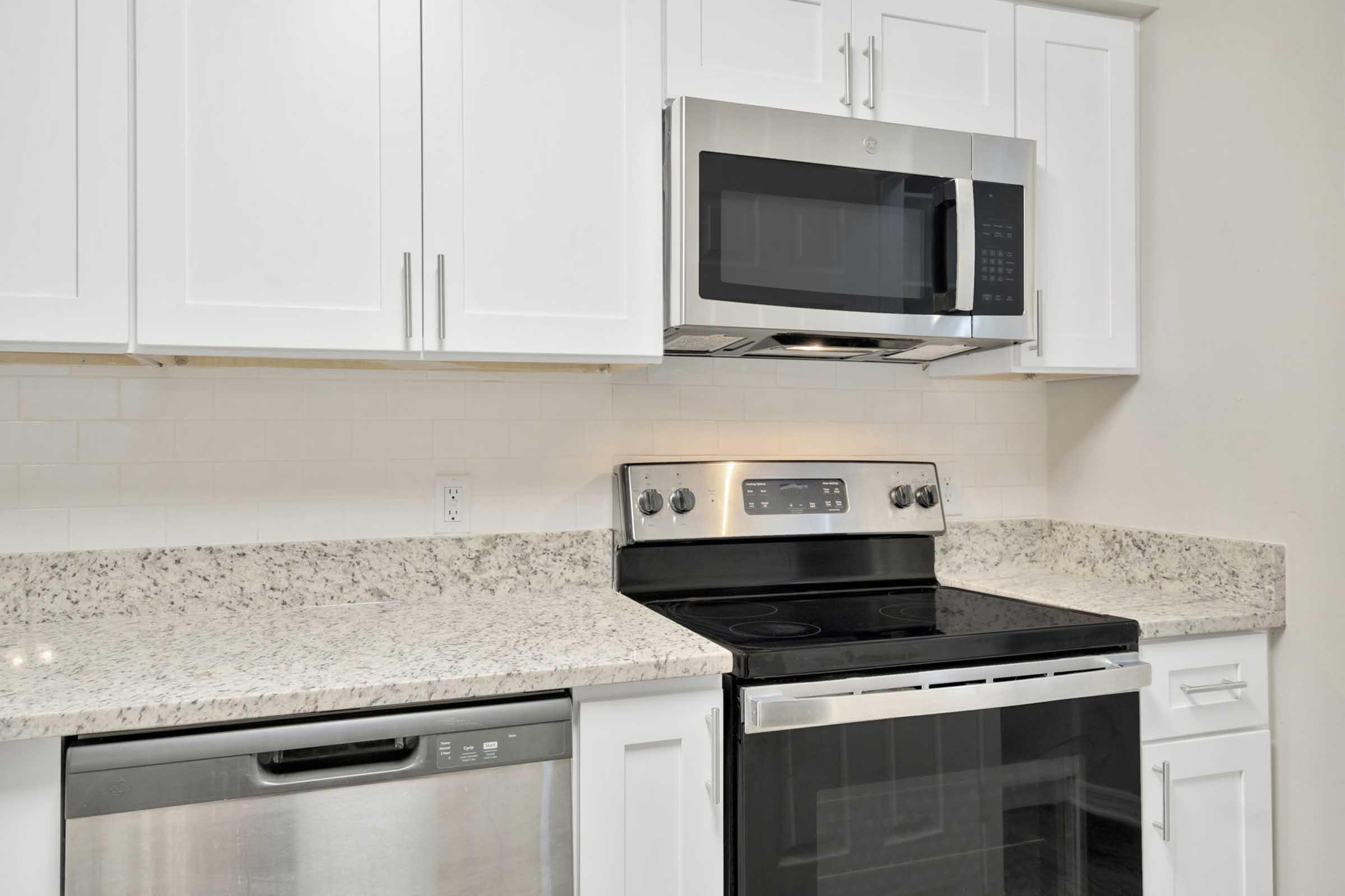 A modern kitchen with white cabinets and a granite countertop. It features a stainless steel microwave above an electric stove and oven, along with a sleek dishwasher. The backsplash is a white tile, creating a clean and vibrant look. Ideal for cooking and entertaining.