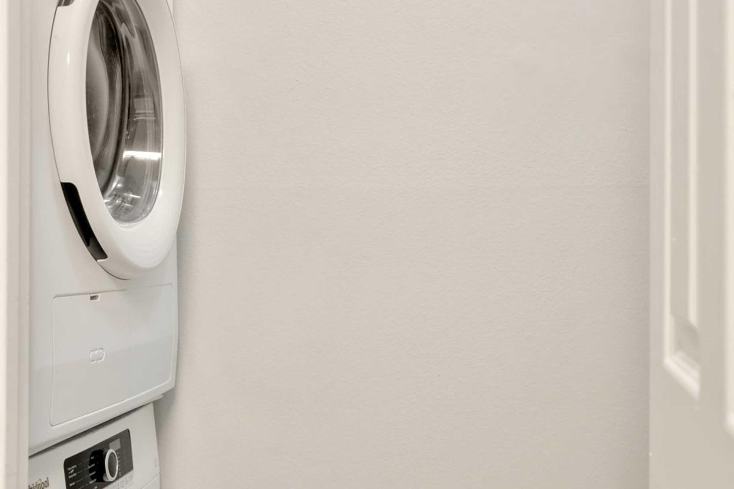 A compact laundry area featuring a white front-loading washer and dryer stacked on top of each other, set against a plain light-colored wall. There is a partially open door to the right, giving a glimpse of the enclosed space.