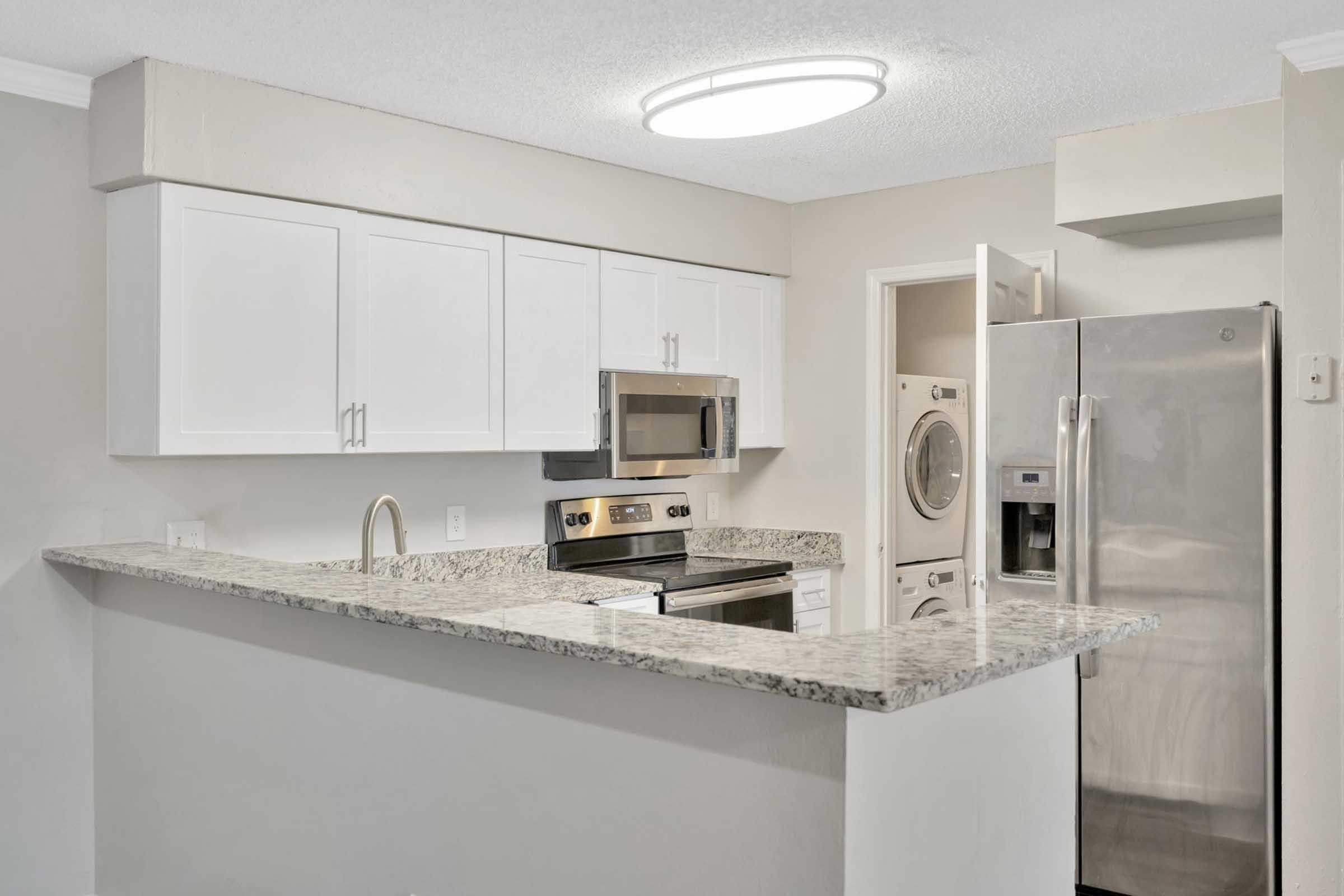 Modern kitchen featuring white cabinets, stainless steel appliances, a granite countertop, and an overhead light fixture. The layout includes a stove, microwave, and refrigerator, with an adjoining space for a washer and dryer visible in the background.