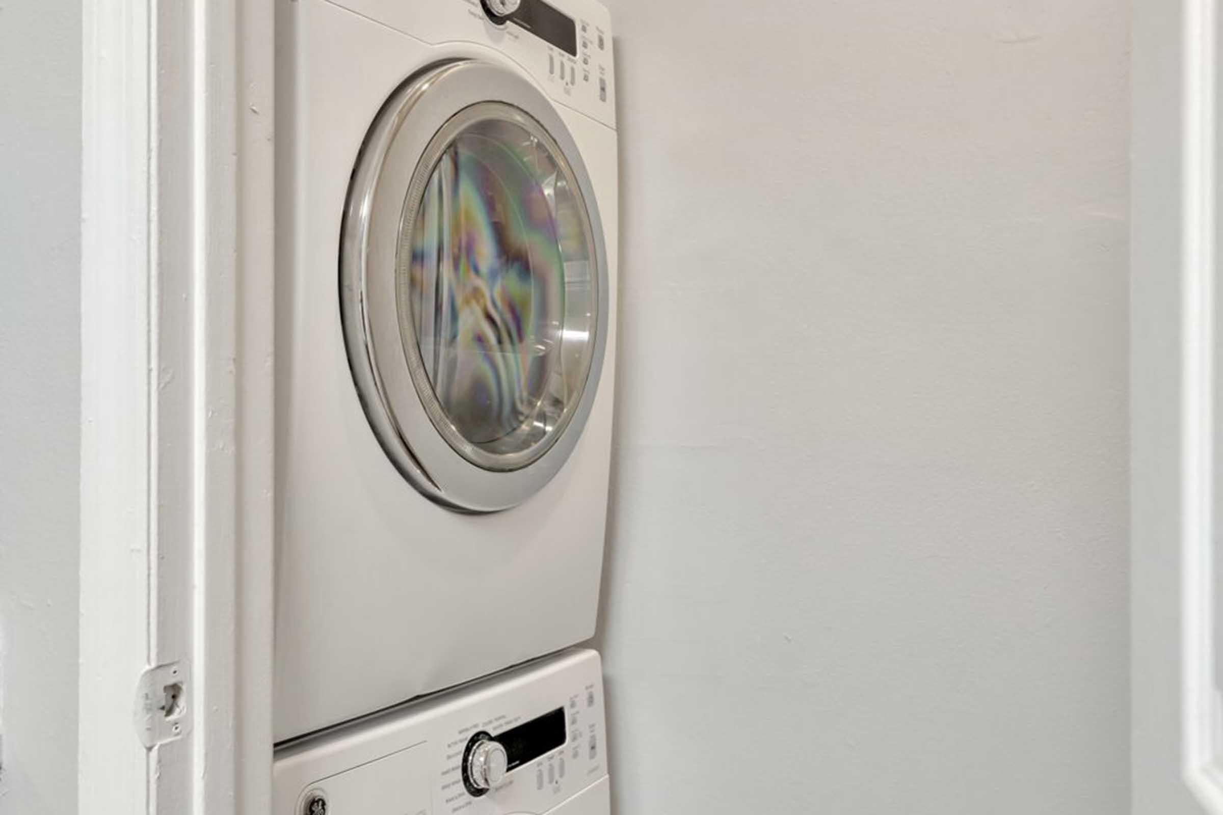 A stacked washer and dryer unit located in a small laundry area against a light gray wall. The washer is on the bottom and the dryer on top, featuring a round glass front on the dryer and control knobs and digital displays on both appliances.