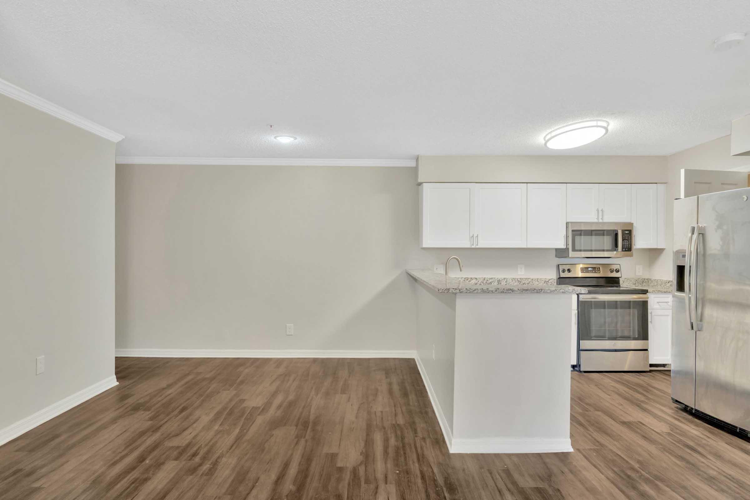 Modern kitchen and living area featuring light-colored walls, laminate flooring, and an open layout. The kitchen has white cabinets, stainless steel appliances, and a spacious countertop. A brightness from ceiling lights enhances the clean and inviting atmosphere.