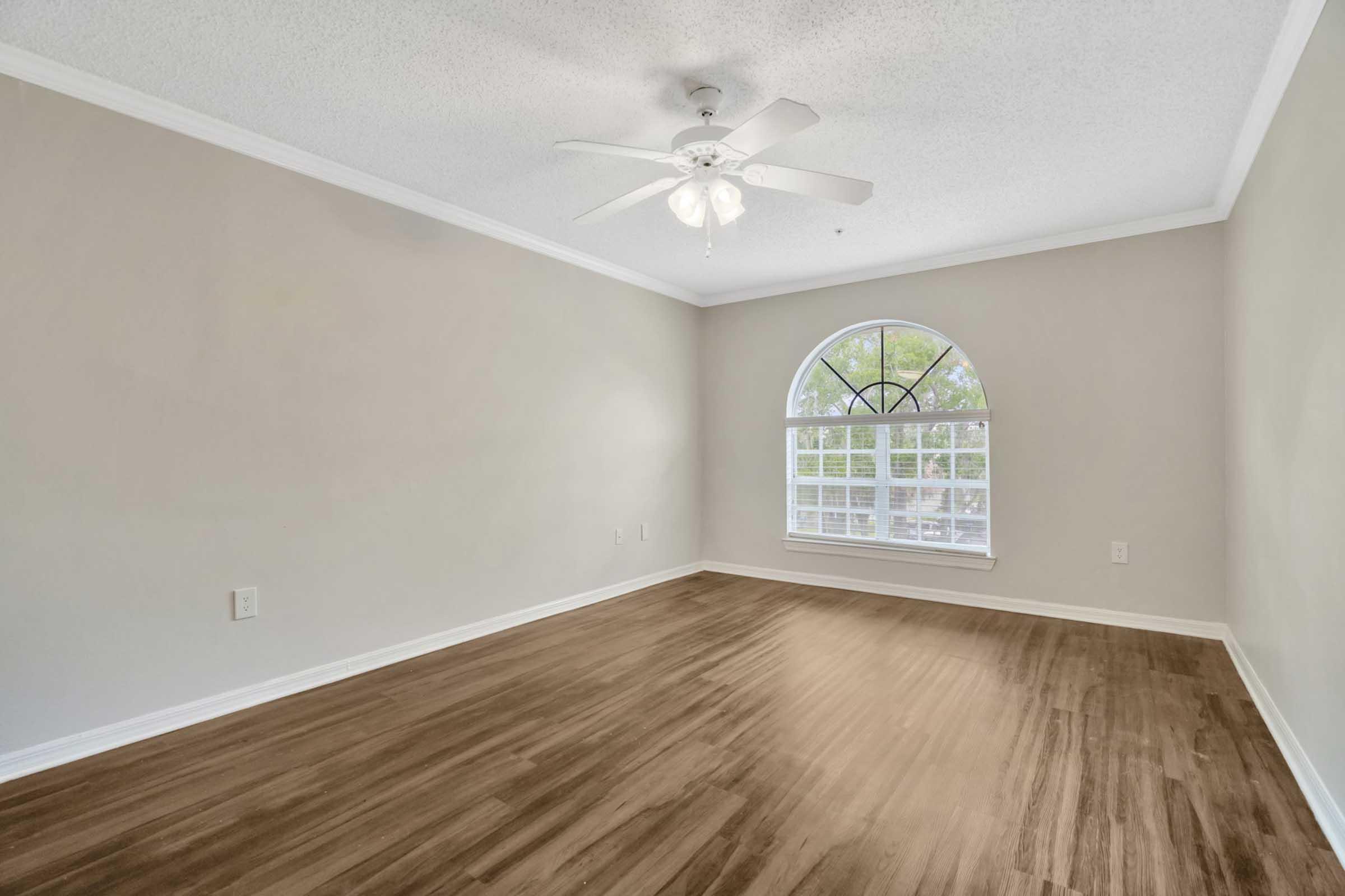 A spacious, empty room featuring a ceiling fan and large arched window allowing natural light. The walls are light-colored, and the floor is made of a warm-toned wood laminate. The room has a serene and clean appearance, ready for personalization or furnishing.