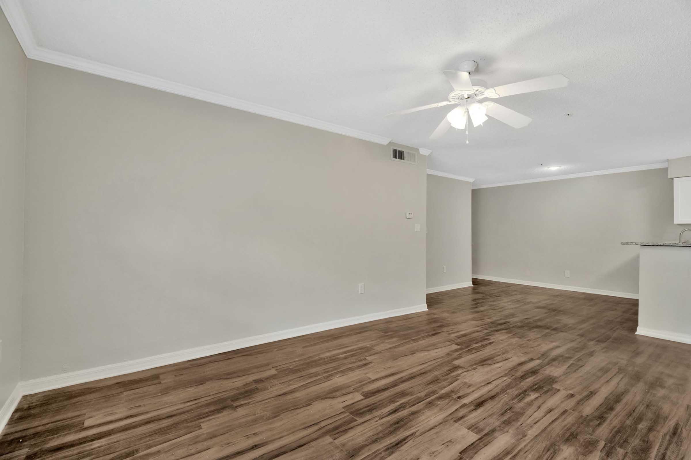 An empty living room with light-colored walls and a ceiling fan. The floor is covered with wooden laminate. There is an open space that leads to a kitchen area in the background, featuring a bar counter. The room is well-lit and appears to be freshly painted with a modern feel.