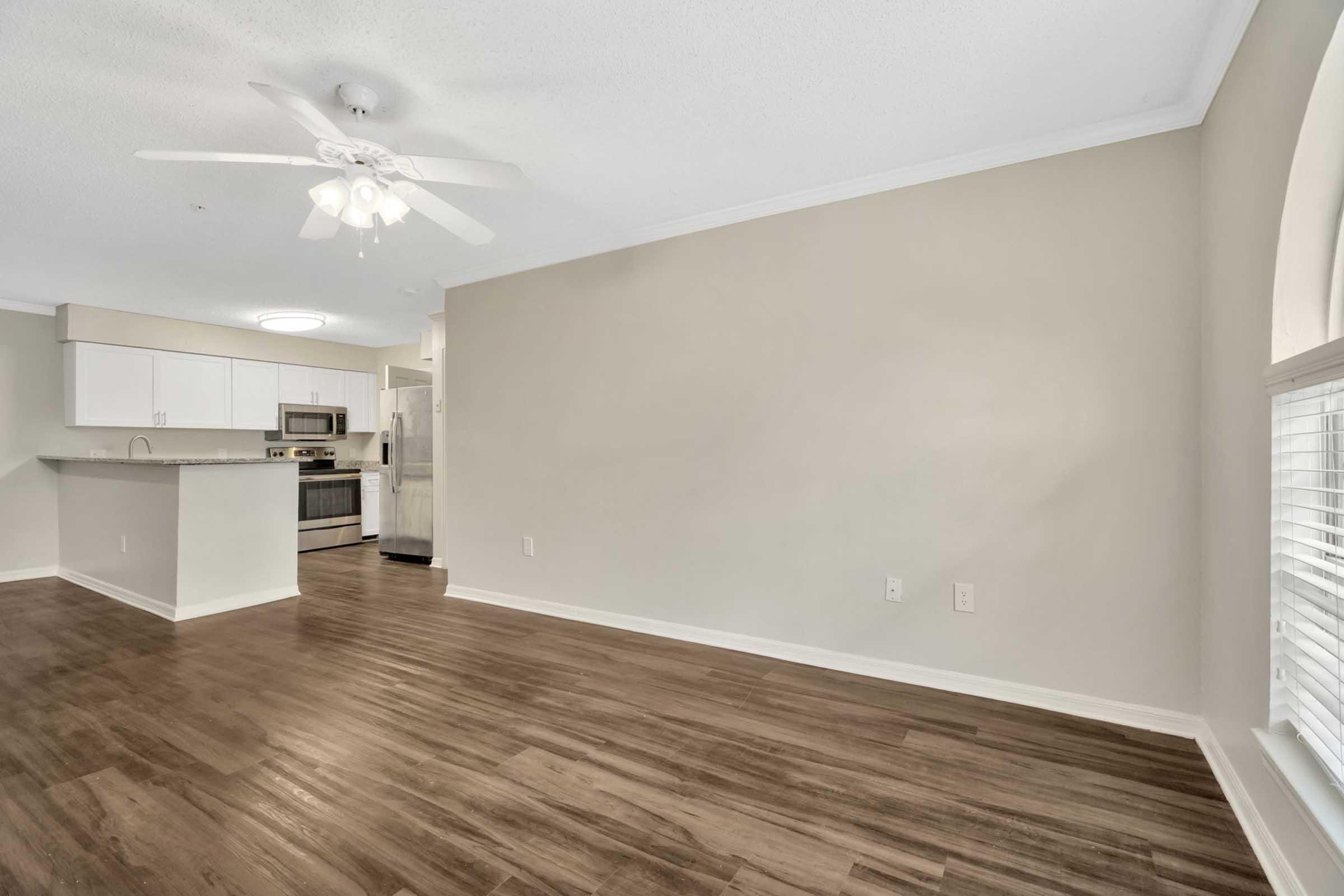 Spacious living area with hardwood floors and light-colored walls. A ceiling fan is visible, and the room opens to a modern kitchen featuring stainless steel appliances. There are large windows allowing natural light, creating a bright and inviting atmosphere.