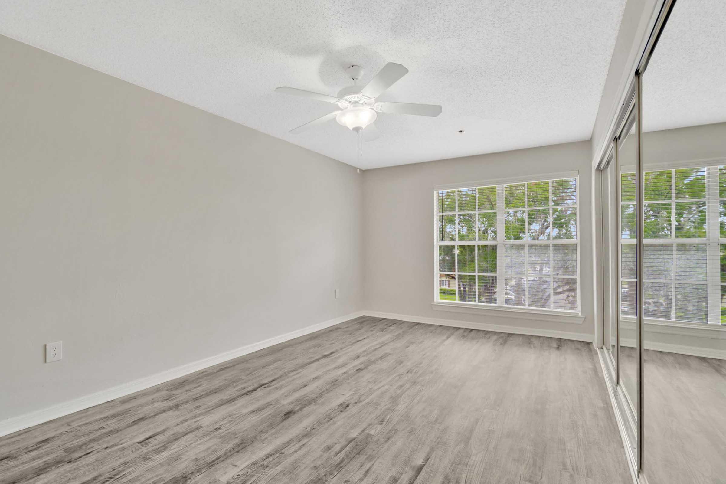 Empty room with light gray walls and a ceiling fan. The floor features light-colored laminate, and there are large windows with blinds allowing natural light. A mirrored closet door is visible on the right side, and the overall space appears clean and minimalist.