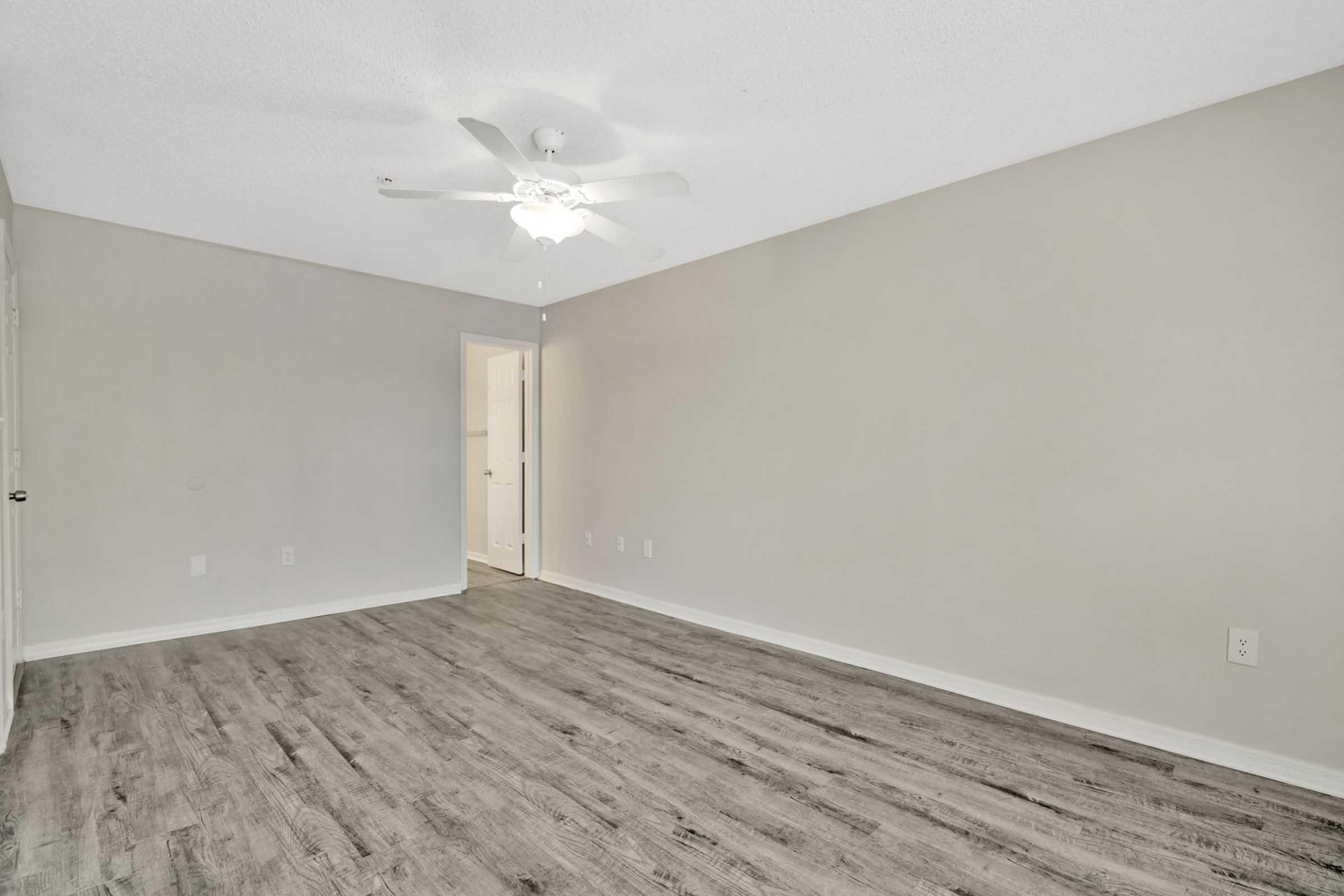 An empty room with light-colored walls and a ceiling fan. The floor is covered with light wood laminate. A doorway is visible on the right side, leading to another space. The room is well-lit, creating a spacious and airy atmosphere.