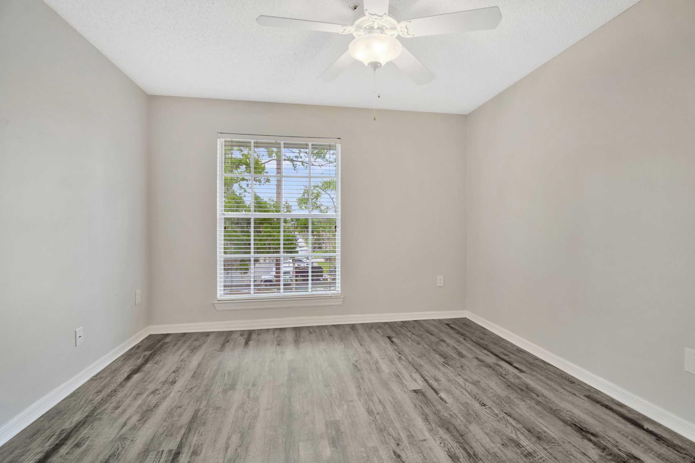 A well-lit empty room featuring light gray walls, a ceiling fan, and a large window with white blinds. The floor is covered with laminate wood flooring. The space appears clean and ready for furnishing.