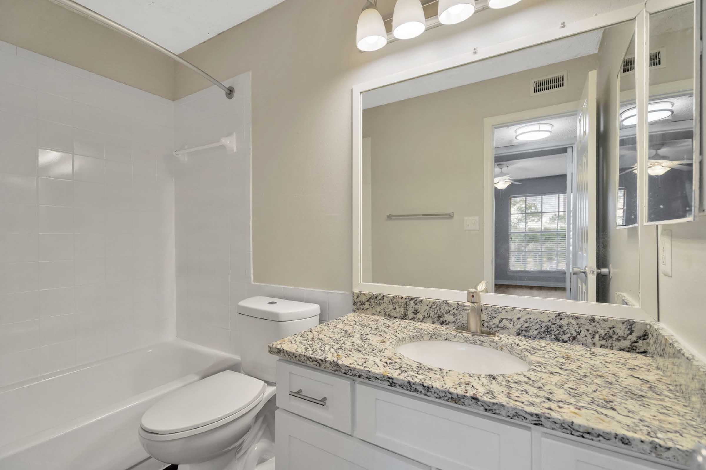 A well-lit bathroom featuring a white bathtub with a shower, a modern sink with a granite countertop, and a large mirror. Natural light streams in from a window in the background, highlighting the contemporary design and neutral color palette. The space is clean and organized, creating a welcoming atmosphere.
