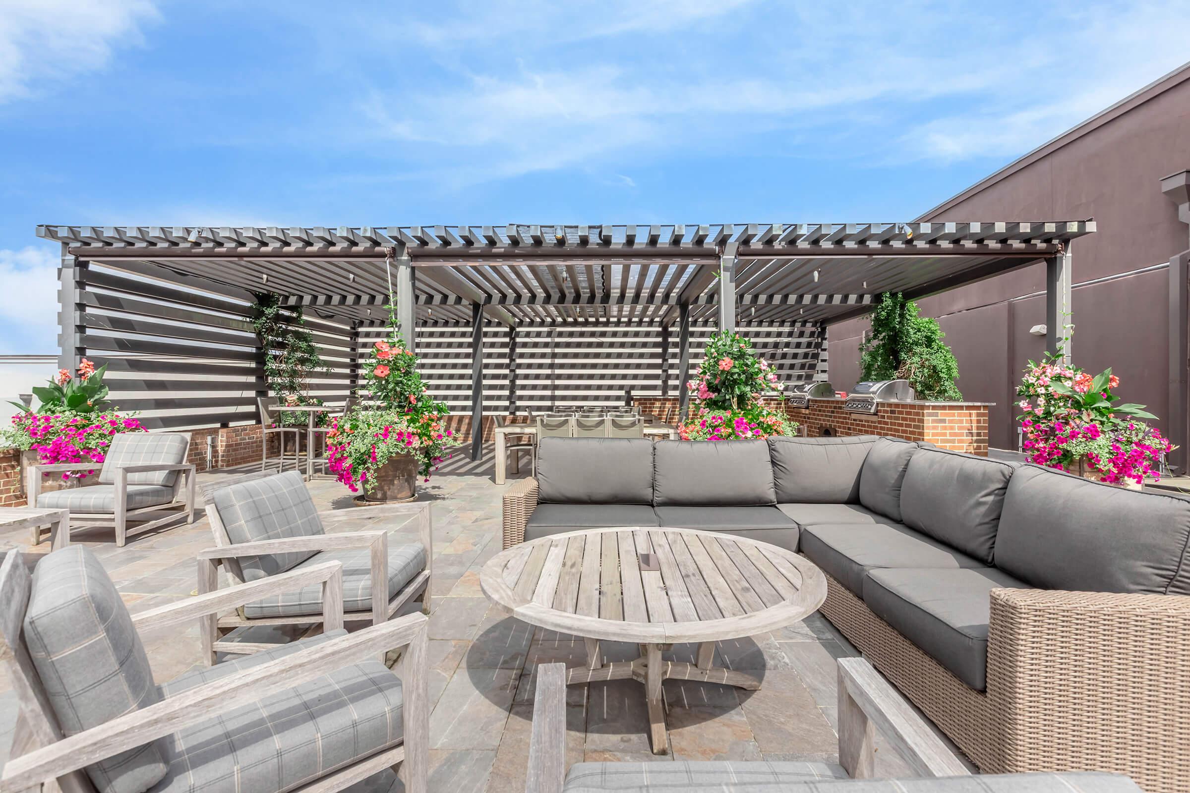 A rooftop terrace featuring a large sectional sofa and a round wooden table. The area is adorned with vibrant flowering plants in pots. A slatted pergola provides partial shade, and the sky is clear with a few clouds, creating a relaxing outdoor space.