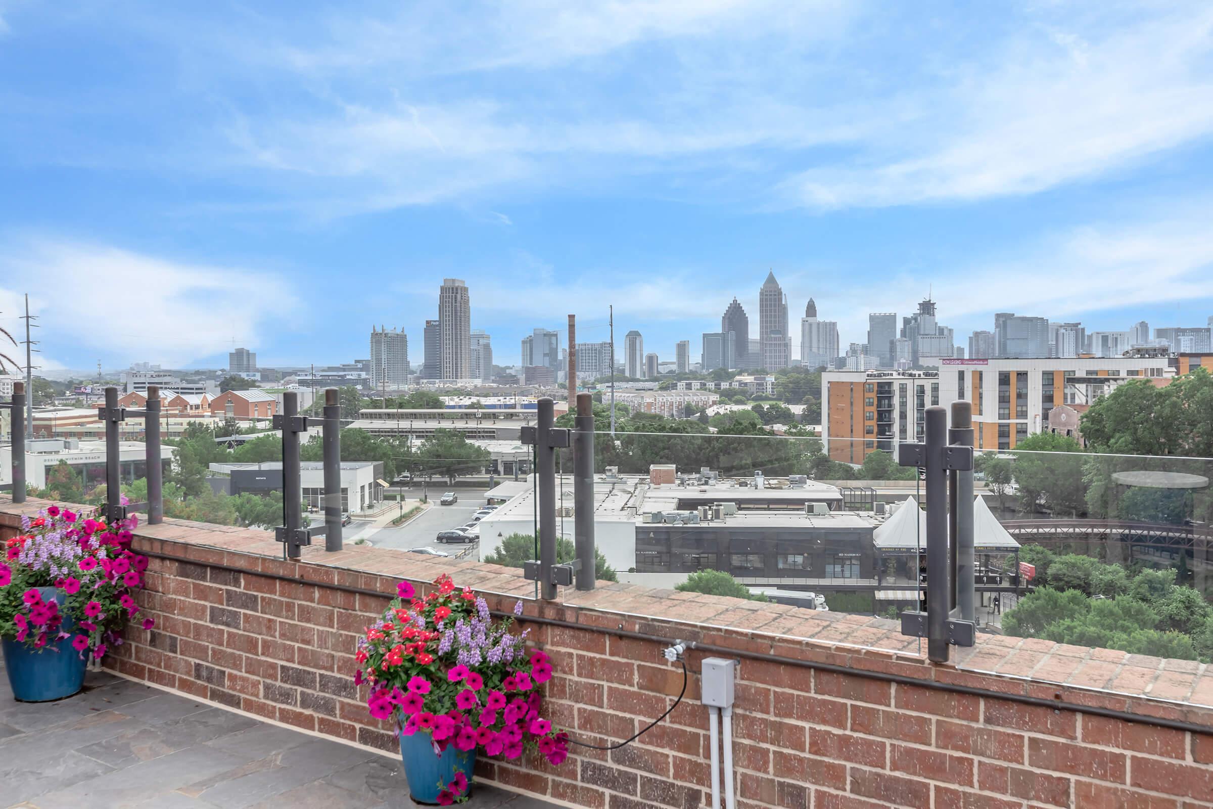 View from a rooftop terrace featuring vibrant flower pots in the foreground and a skyline of a city in the background. The scene includes modern buildings under a clear blue sky.