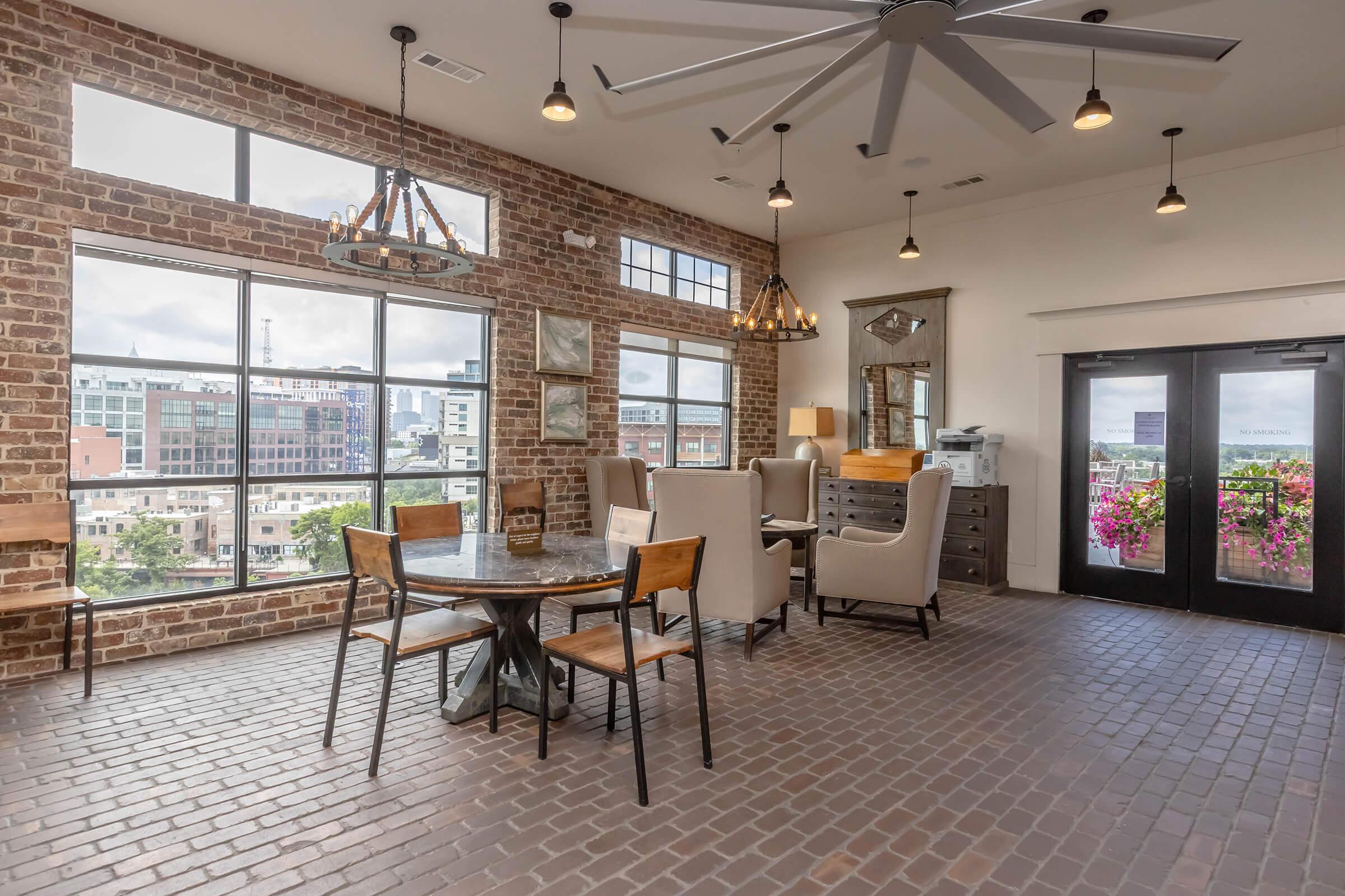 A spacious, well-lit interior featuring exposed brick walls and large windows. There is a round table with four wooden chairs in the center, surrounded by stylish armchairs. Ceiling fans hang above, and there are decorative light fixtures. The entrance has double doors leading outside, framed by colorful flowers.