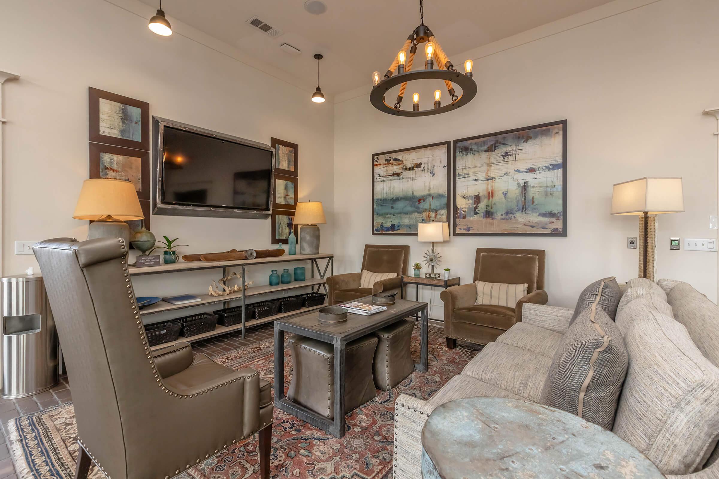 A cozy living room featuring two armchairs, a sofa, and a coffee table. The walls are adorned with decorative artwork, and there is a flat-screen TV mounted on the wall. Two lamps provide warm lighting, and a chandelier hangs from the ceiling. The room has a welcoming, modern aesthetic with earthy tones and a rug on the floor.
