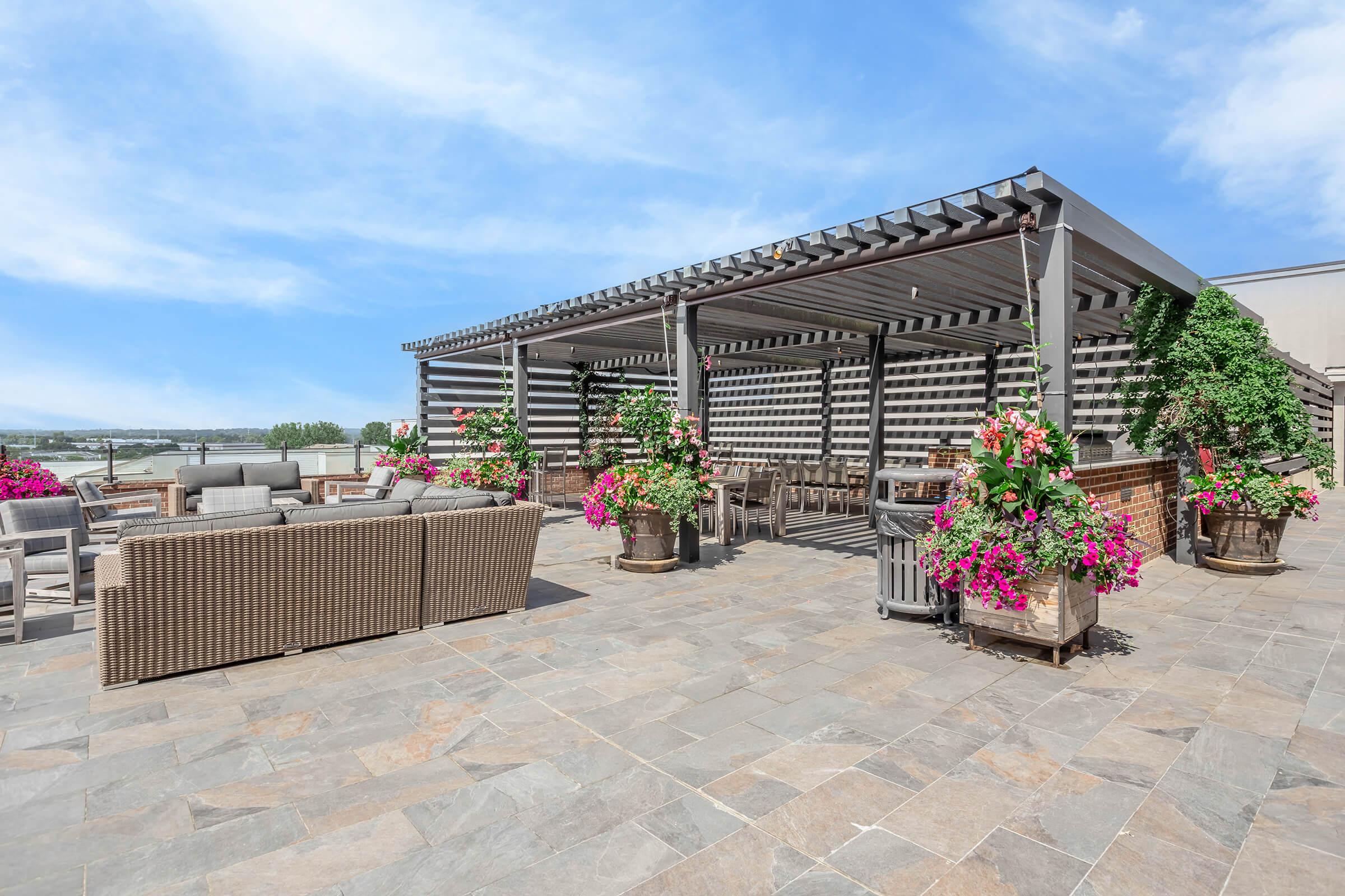 A spacious rooftop patio featuring a shaded seating area with comfortable furniture, surrounded by vibrant flowerpots. The stone floor adds an elegant touch, while the structured awning provides shelter. The backdrop includes a clear blue sky and distant greenery, creating a relaxing outdoor environment.
