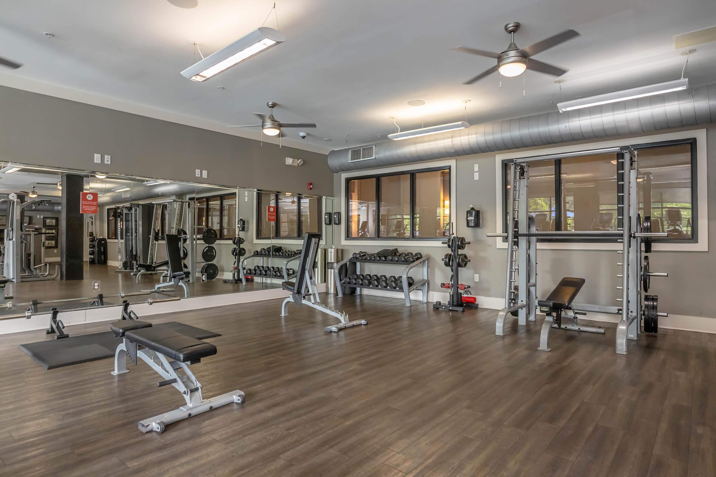 A modern gym interior featuring a variety of workout equipment, including weight benches, a weightlifting machine, and dumbbells organized on racks. The space has large mirrors, ceiling fans, and a well-lit environment, creating a functional and inviting atmosphere for exercise.
