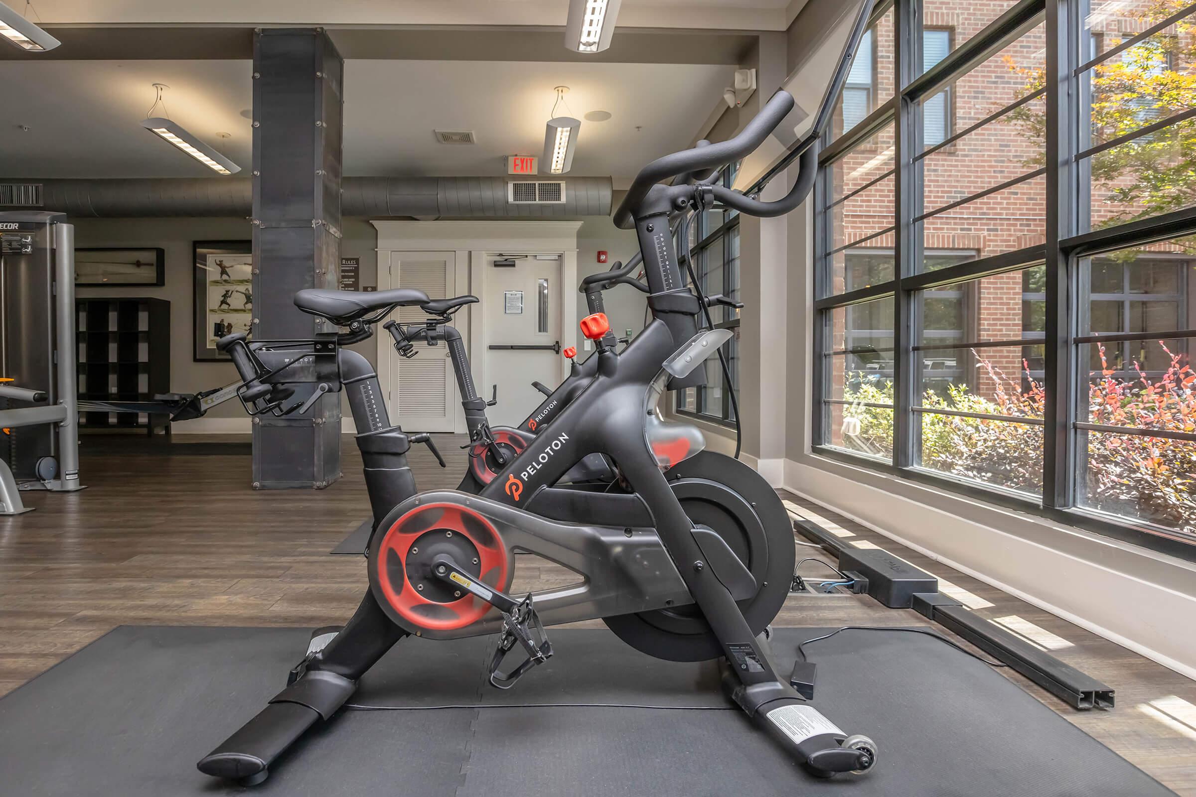 A stationary exercise bike positioned in a modern gym setting, featuring large windows that let in natural light. The bike is sleek and black with red accents, situated on a rubber mat. The background includes gym equipment and a view of greenery outside.