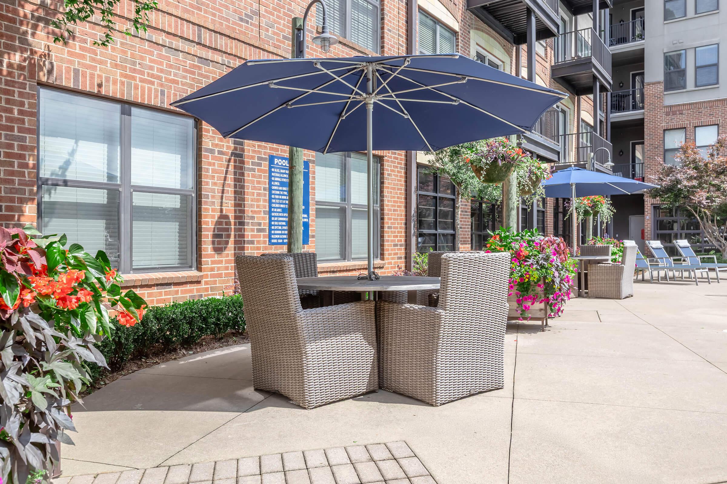 A cozy outdoor patio area featuring a table with four wicker chairs underneath a large navy umbrella. Surrounding the space are colorful flower arrangements and additional lounge chairs, with a backdrop of modern apartment buildings and large windows.